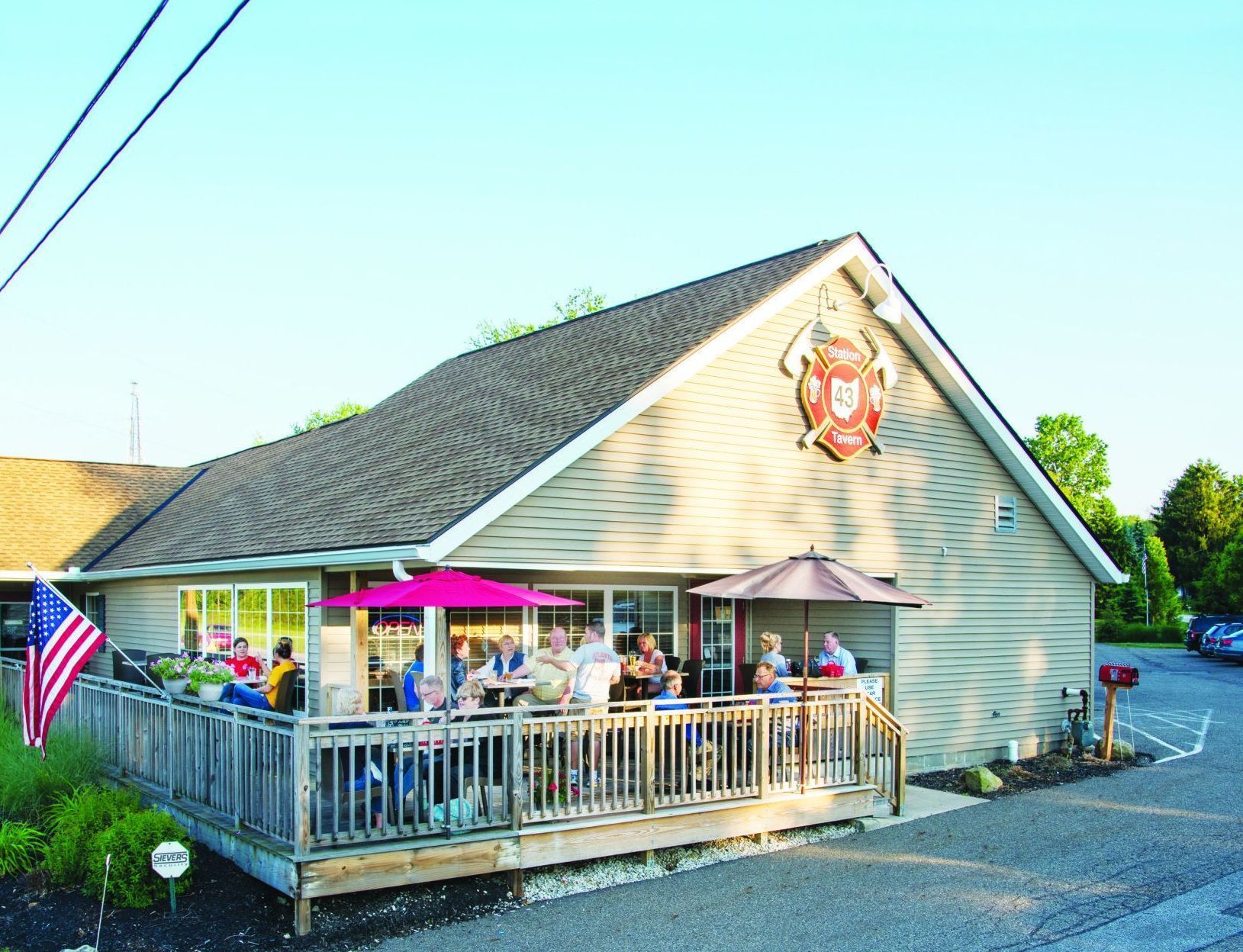 Outdoor patio of a restaurant with people seated at tables, red umbrellas, American flag.