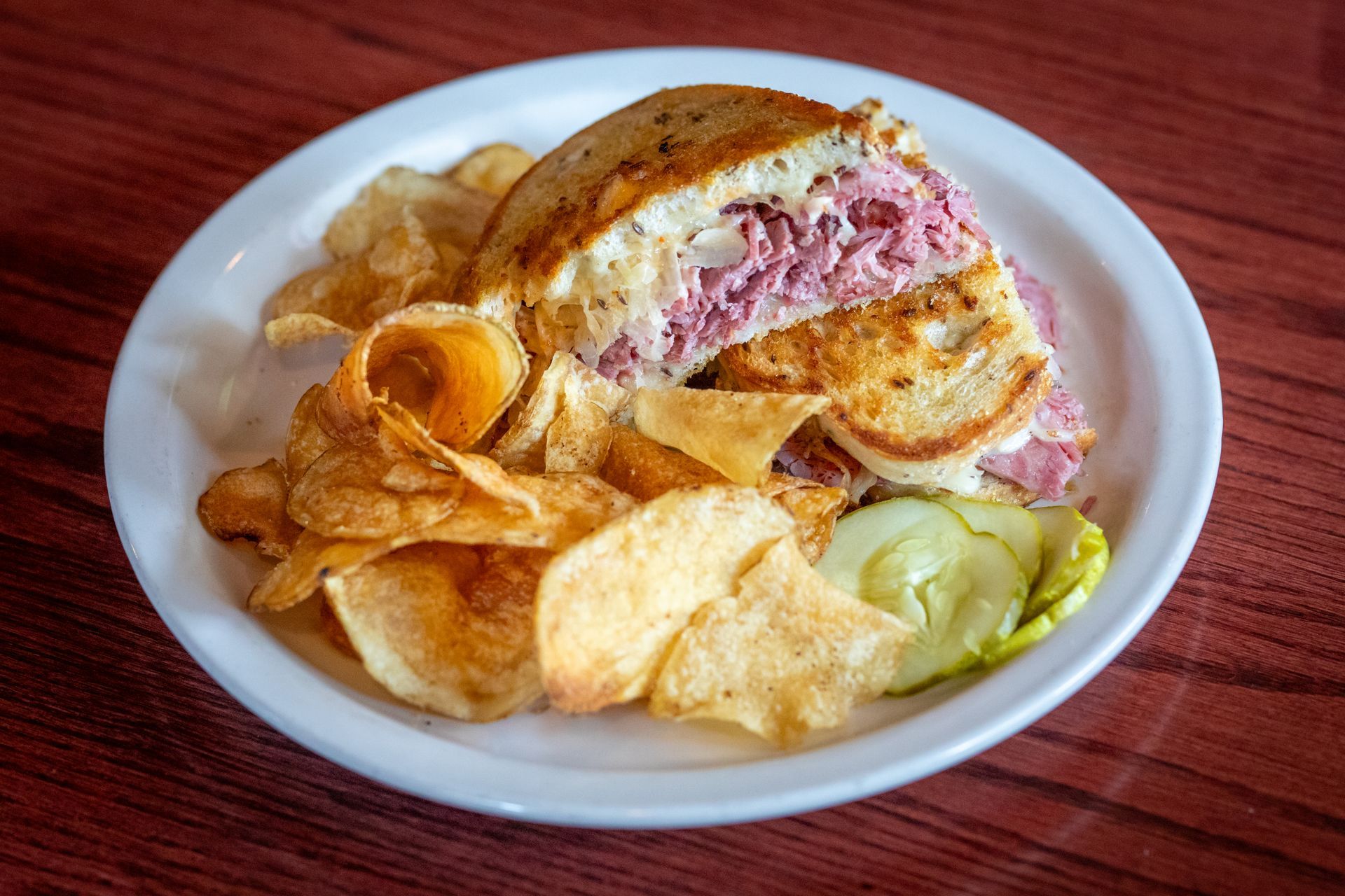 Reuben sandwich with potato chips and a pickle on a white plate, wood table.