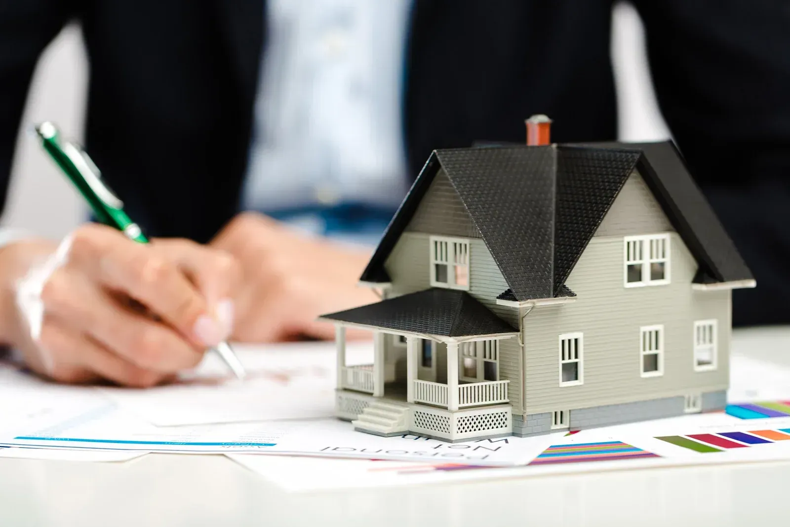 Miniature house on table; person in suit writing on documents.