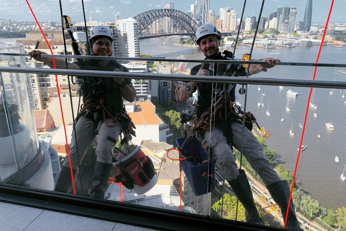 Two window washers suspended outside a high-rise with the Harbour Bridge in the background