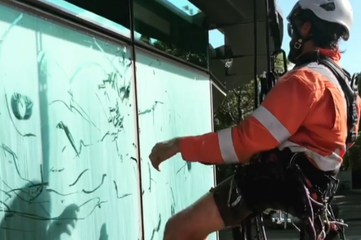 Person in orange climbing gear rappels beside a glass wall with cracks and rope reflections.