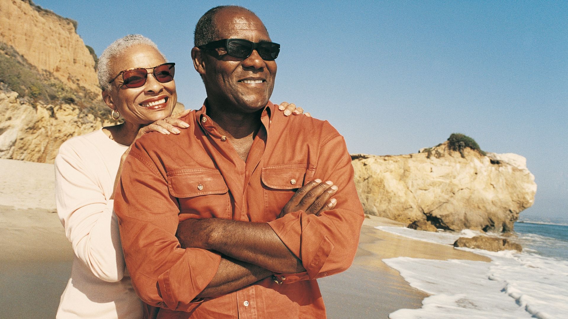 A man and a woman are standing on a beach with their arms crossed.