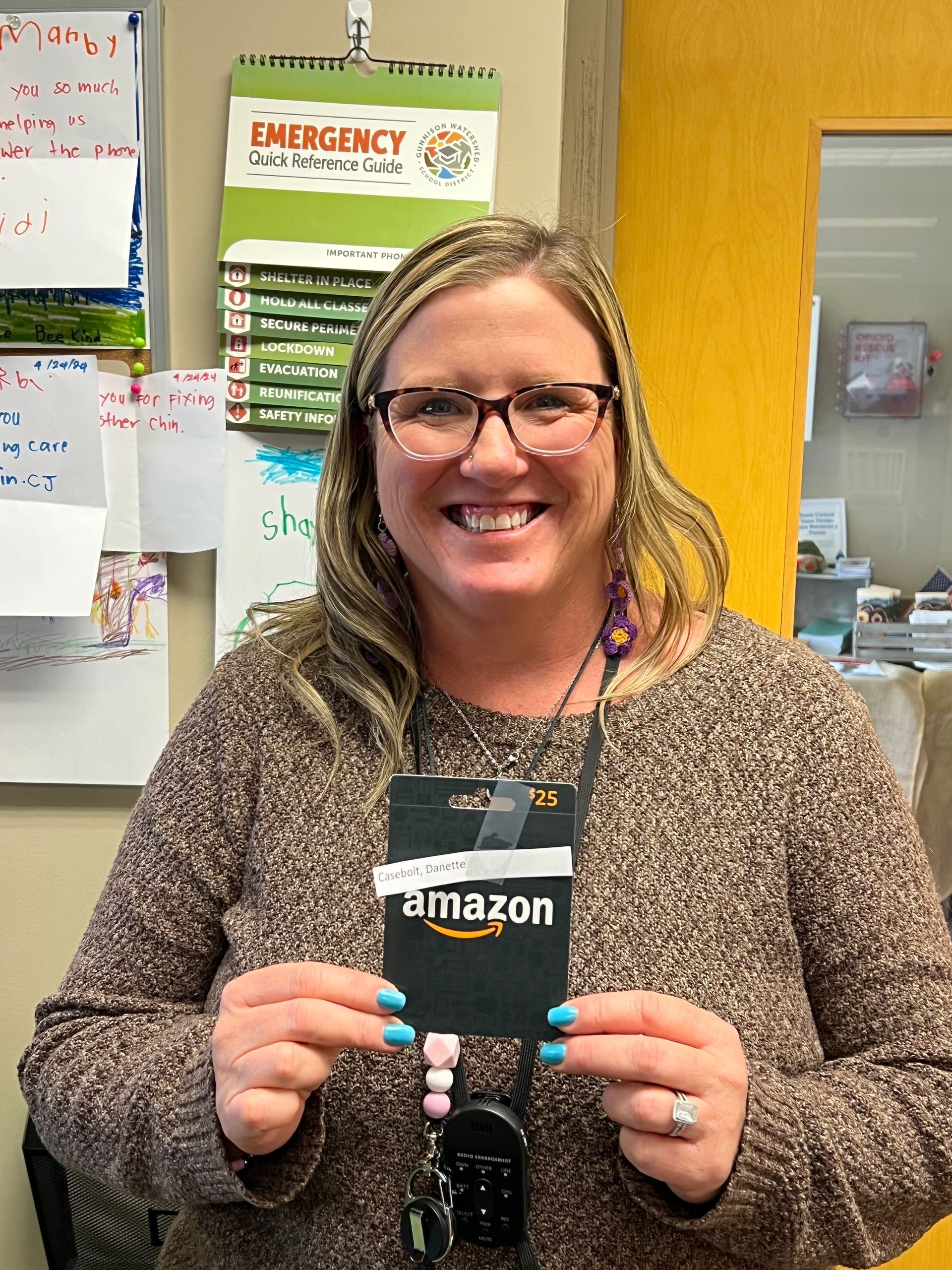 Woman smiling, holding an Amazon gift card. Brown sweater, glasses. Office setting.