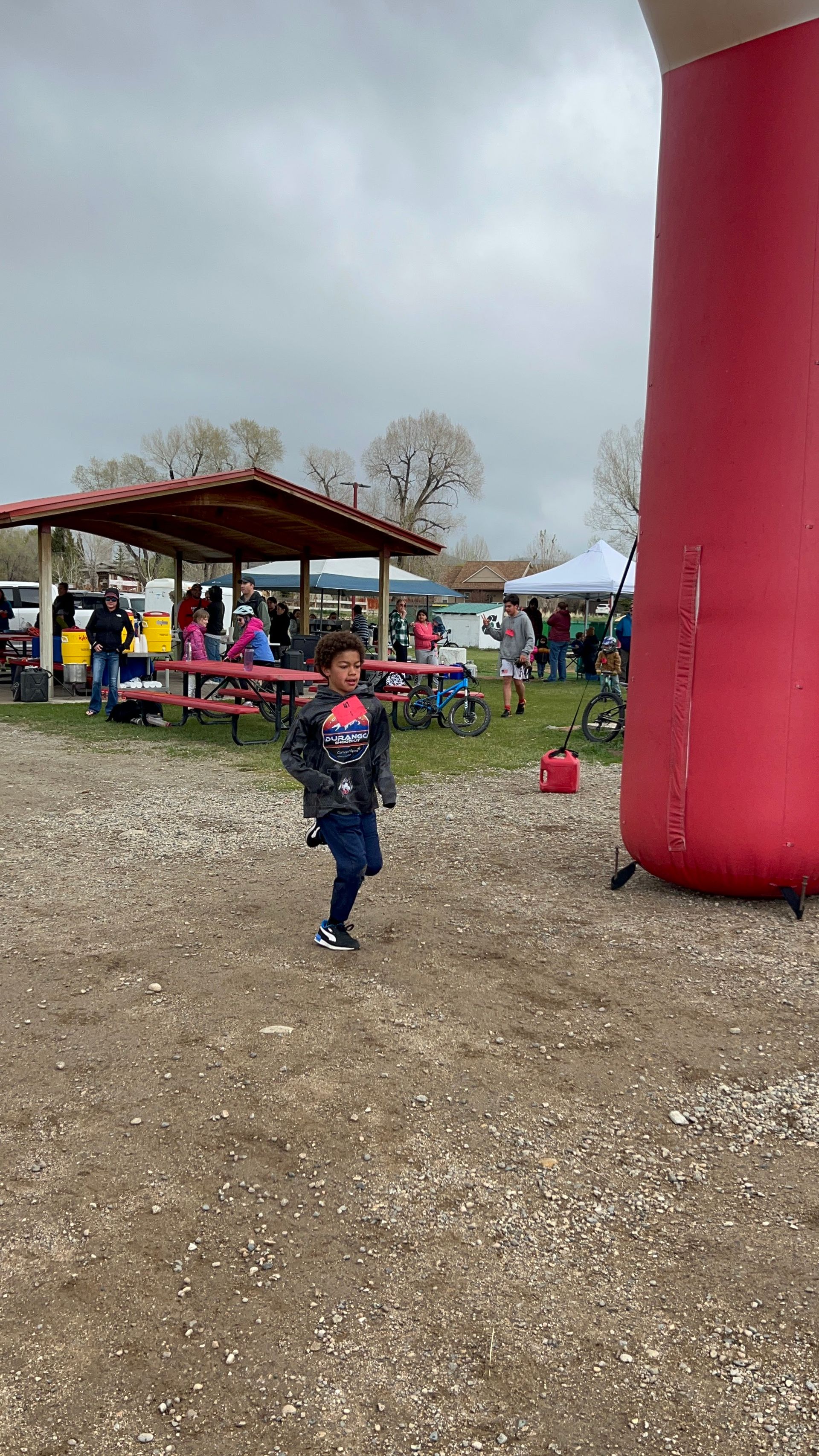 Child running, wearing jacket, near red inflatable arch, and picnic area. Cloudy day.