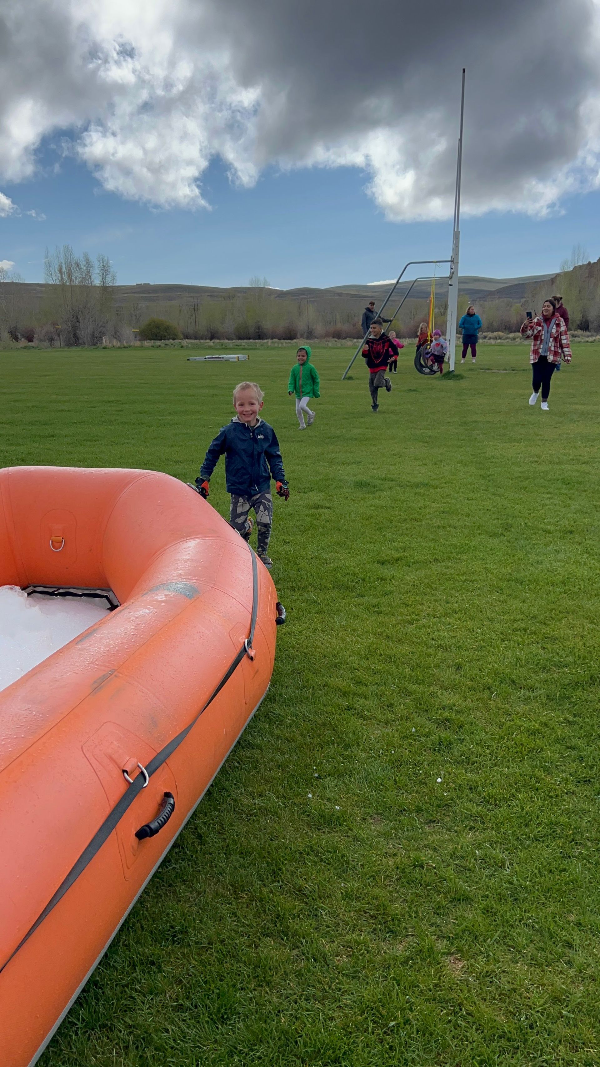 Children running on a grassy field; an orange inflatable boat in the foreground. Bright sunny day, cloudy sky.