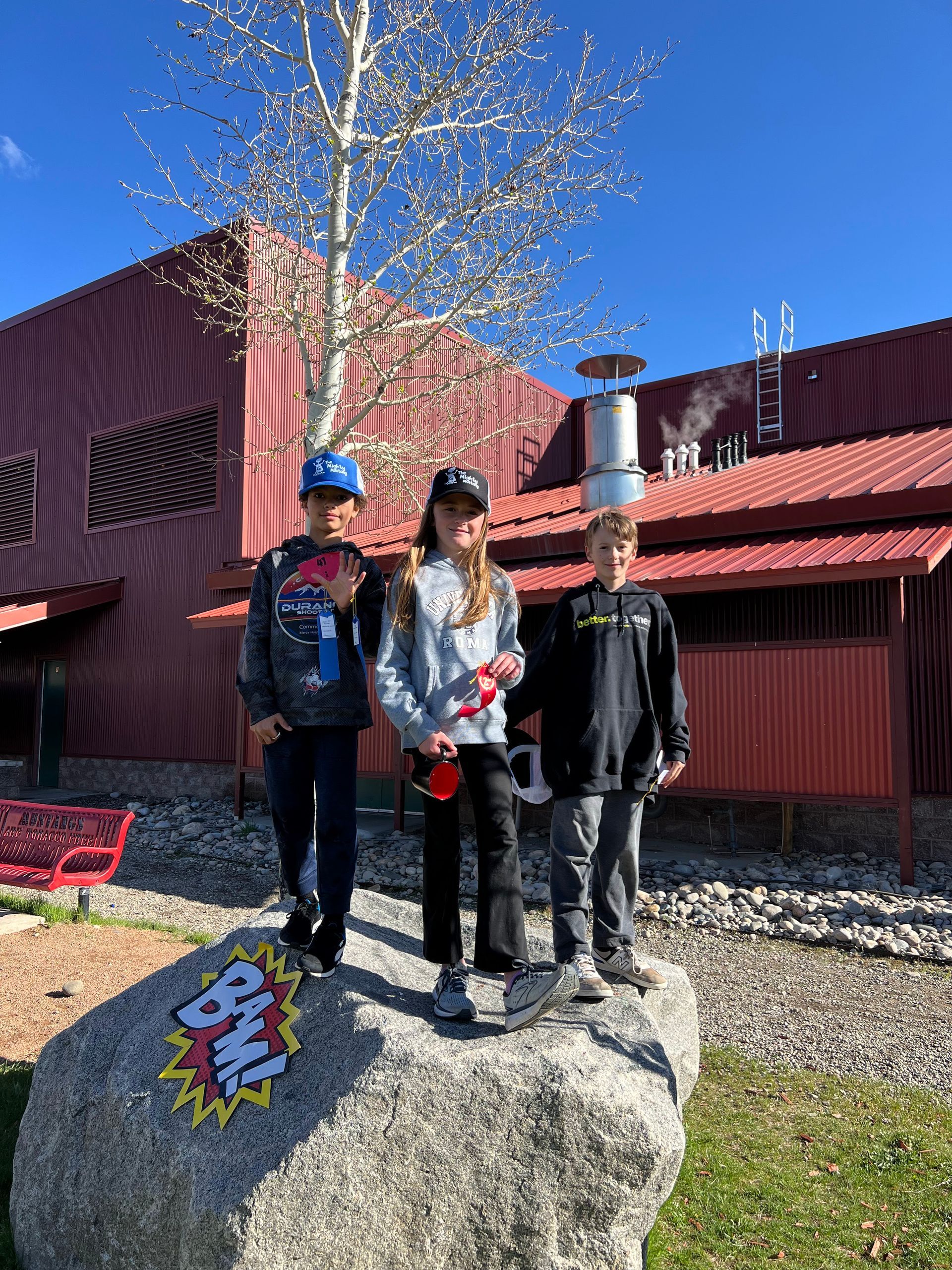 Three children standing on a rock in front of a red building, under a sunny sky.