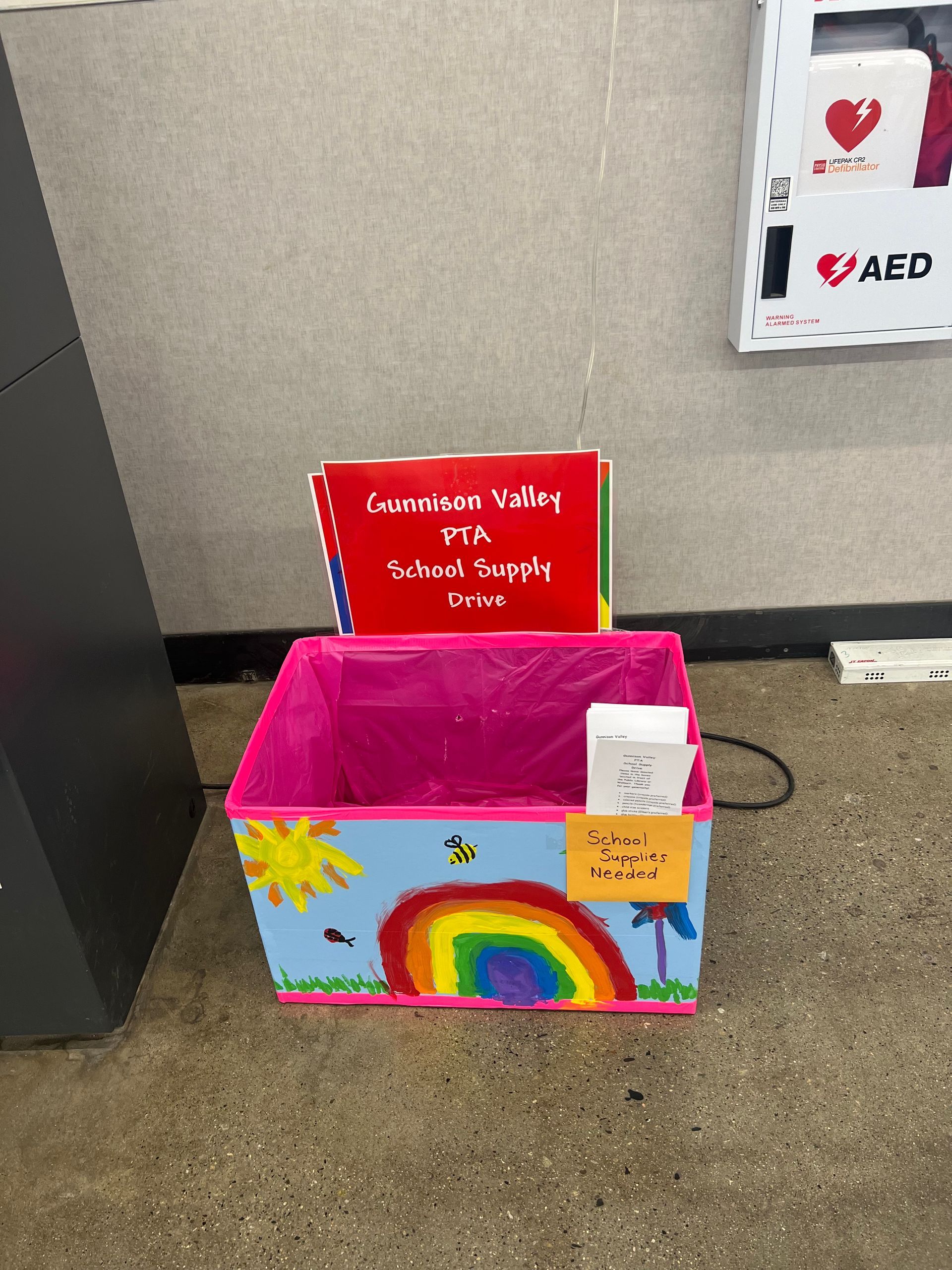 A decorated box with books inside, near an AED and a black object. The box has a rainbow and a sun drawn on it.
