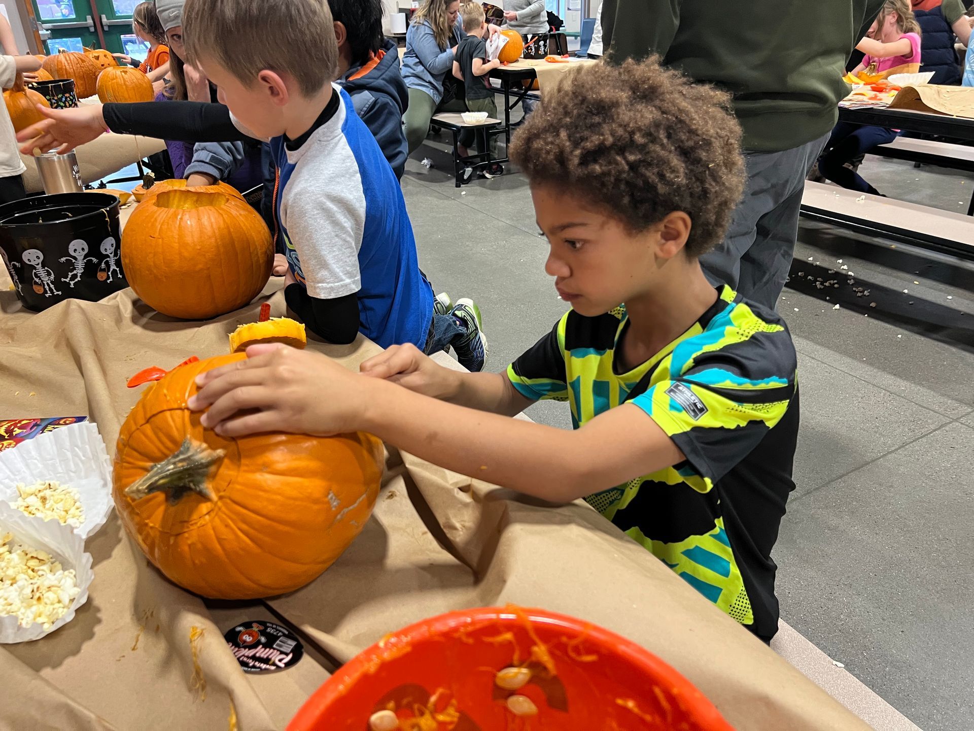 Boy in colorful shirt carving a pumpkin at a table with other children during a fall activity.