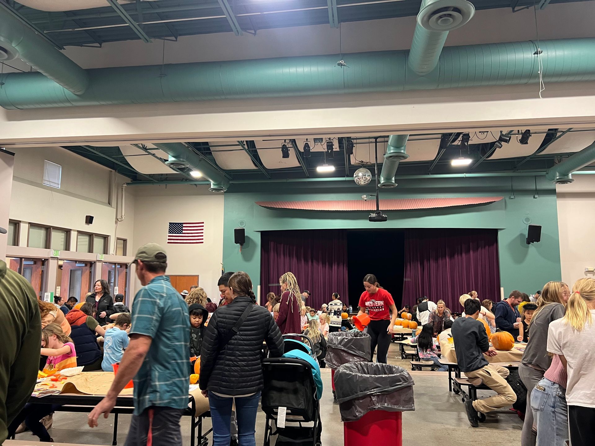 People at tables in a large room, possibly an event. Some are carving pumpkins, there's a stage with maroon curtains.