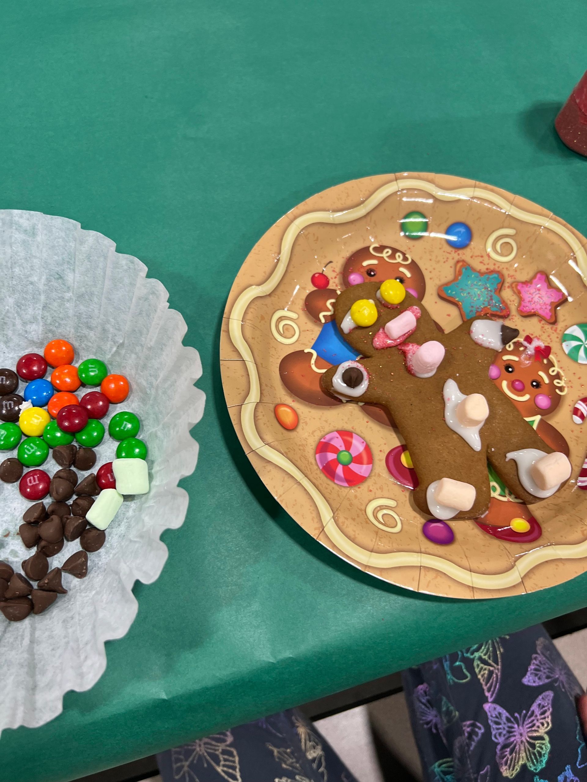 Gingerbread man and candies on a plate with toppings, alongside assorted candies in a paper cup, all on a green surface.