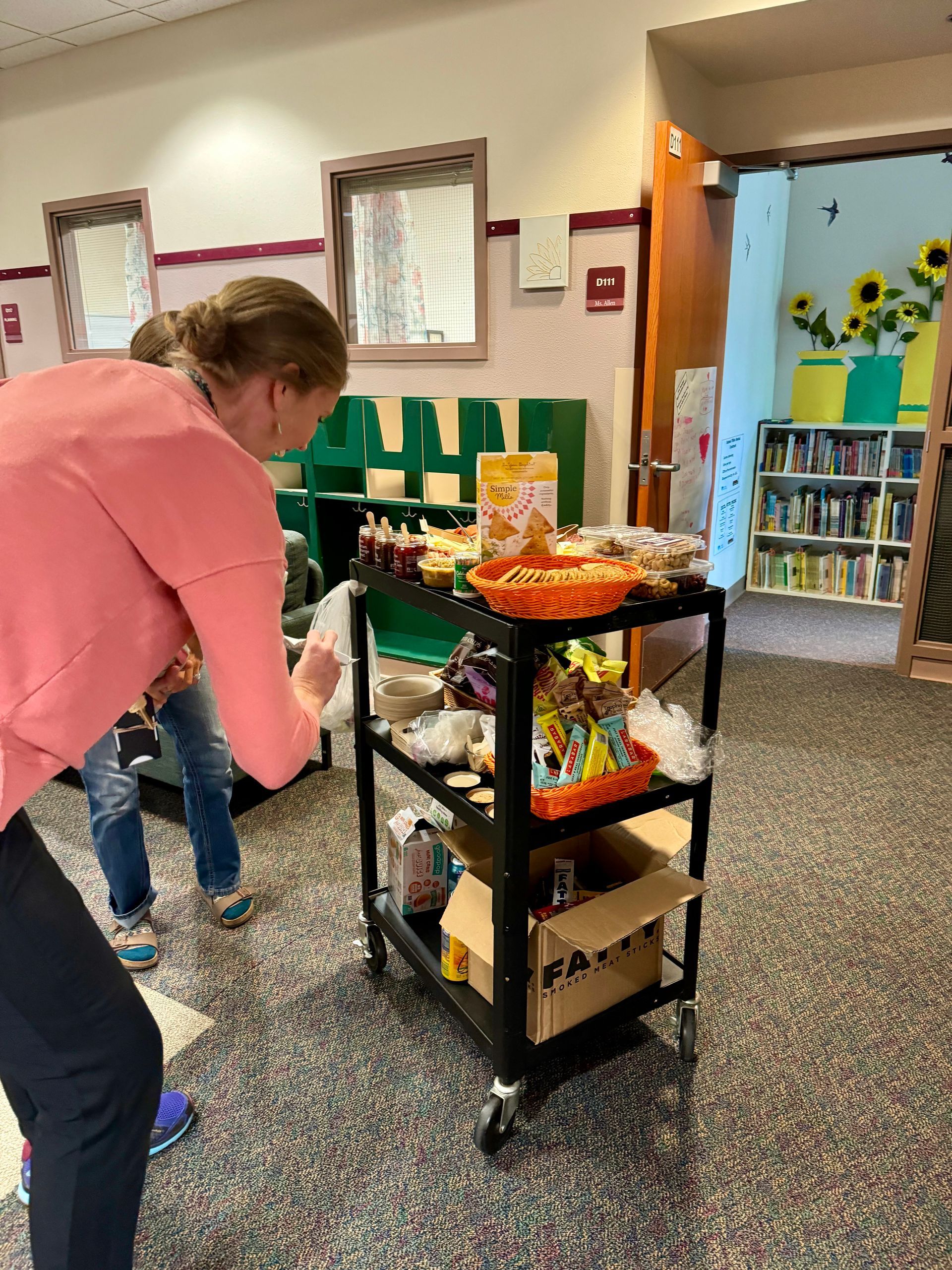 Woman and child at a cart with snacks in a hallway; the woman is reaching toward the items on the cart.