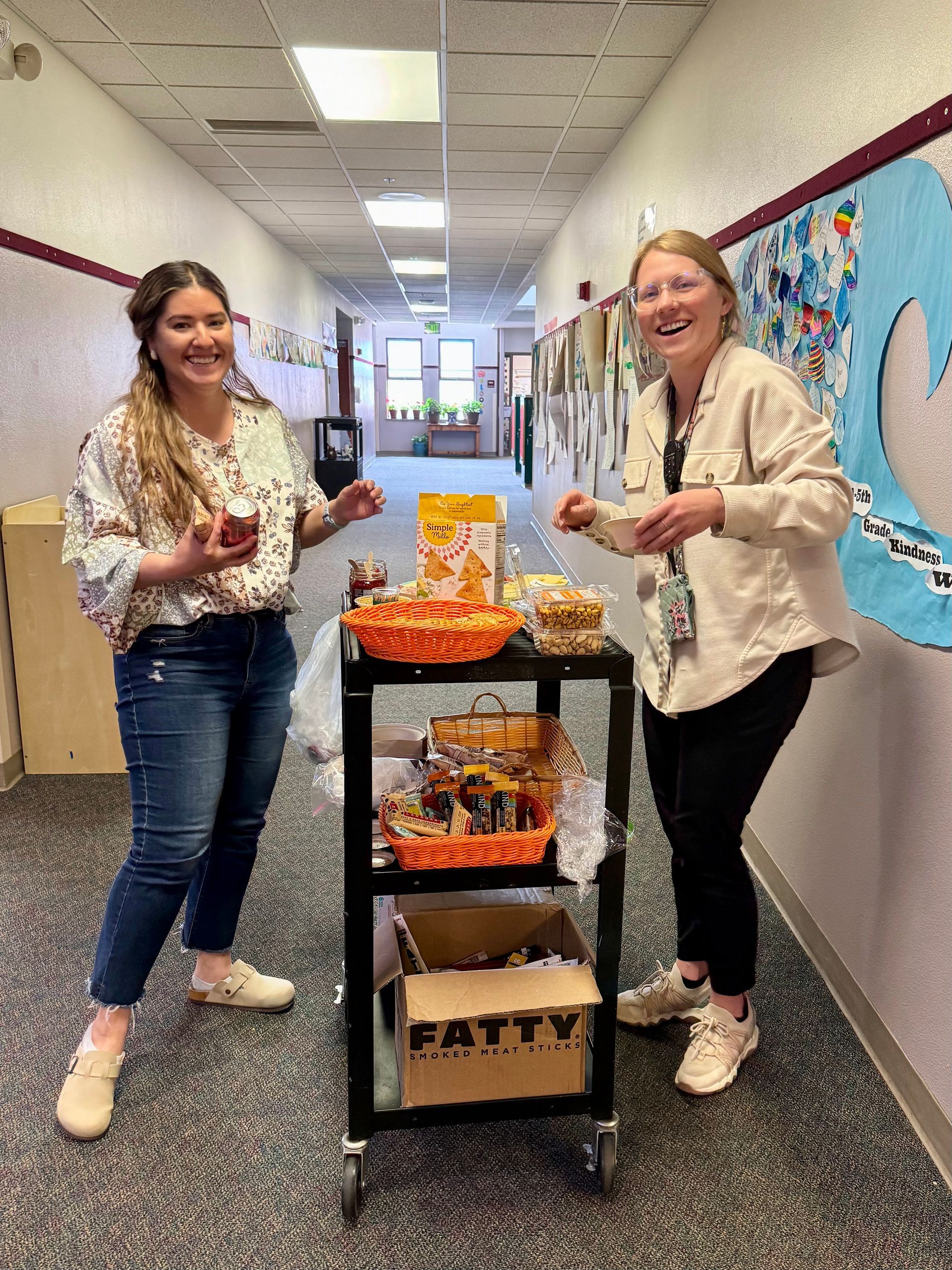 Two people smile beside a cart of snacks in a school hallway.