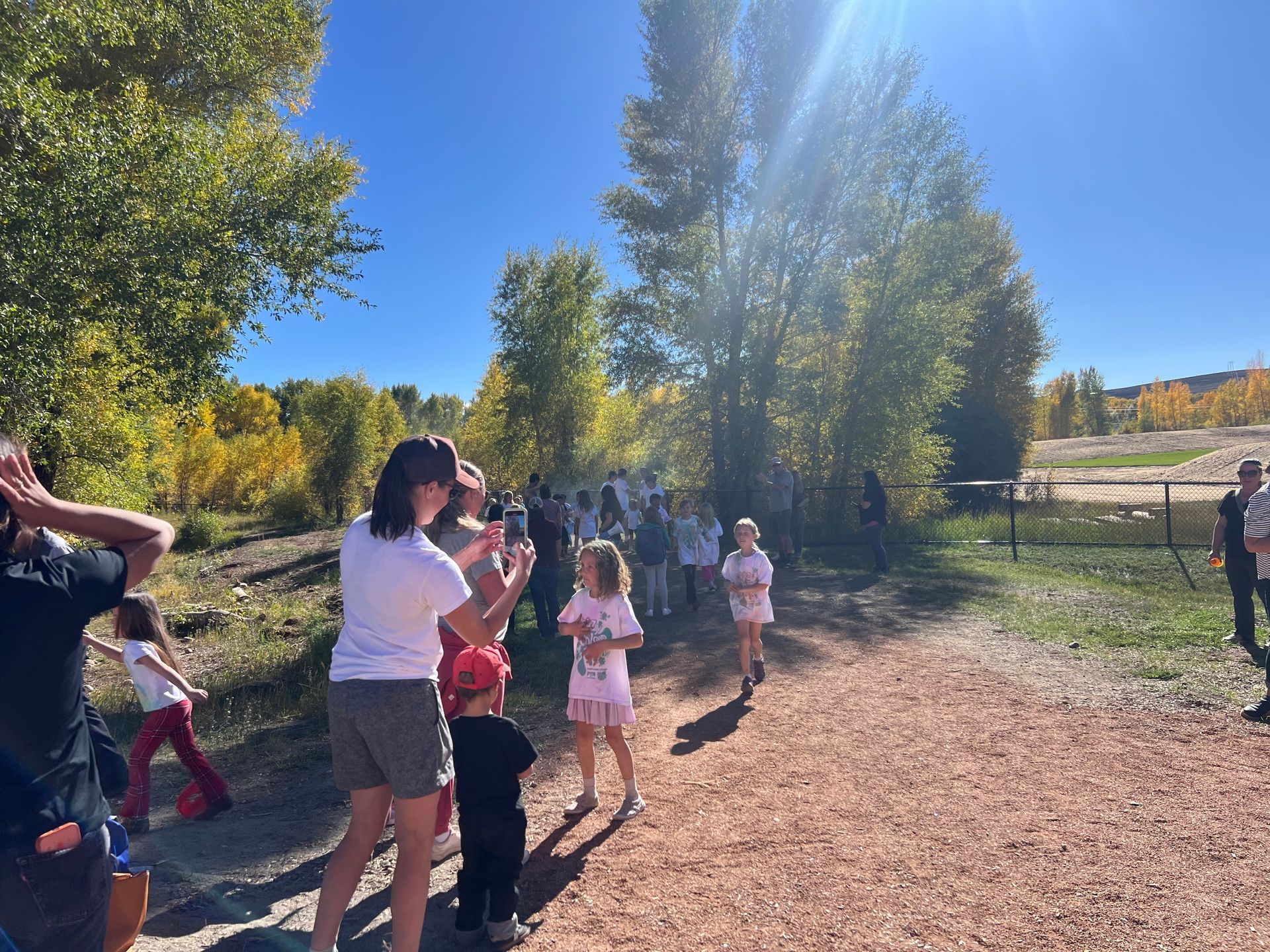People watch children running on a dirt path in autumn, trees with yellow leaves in the background.