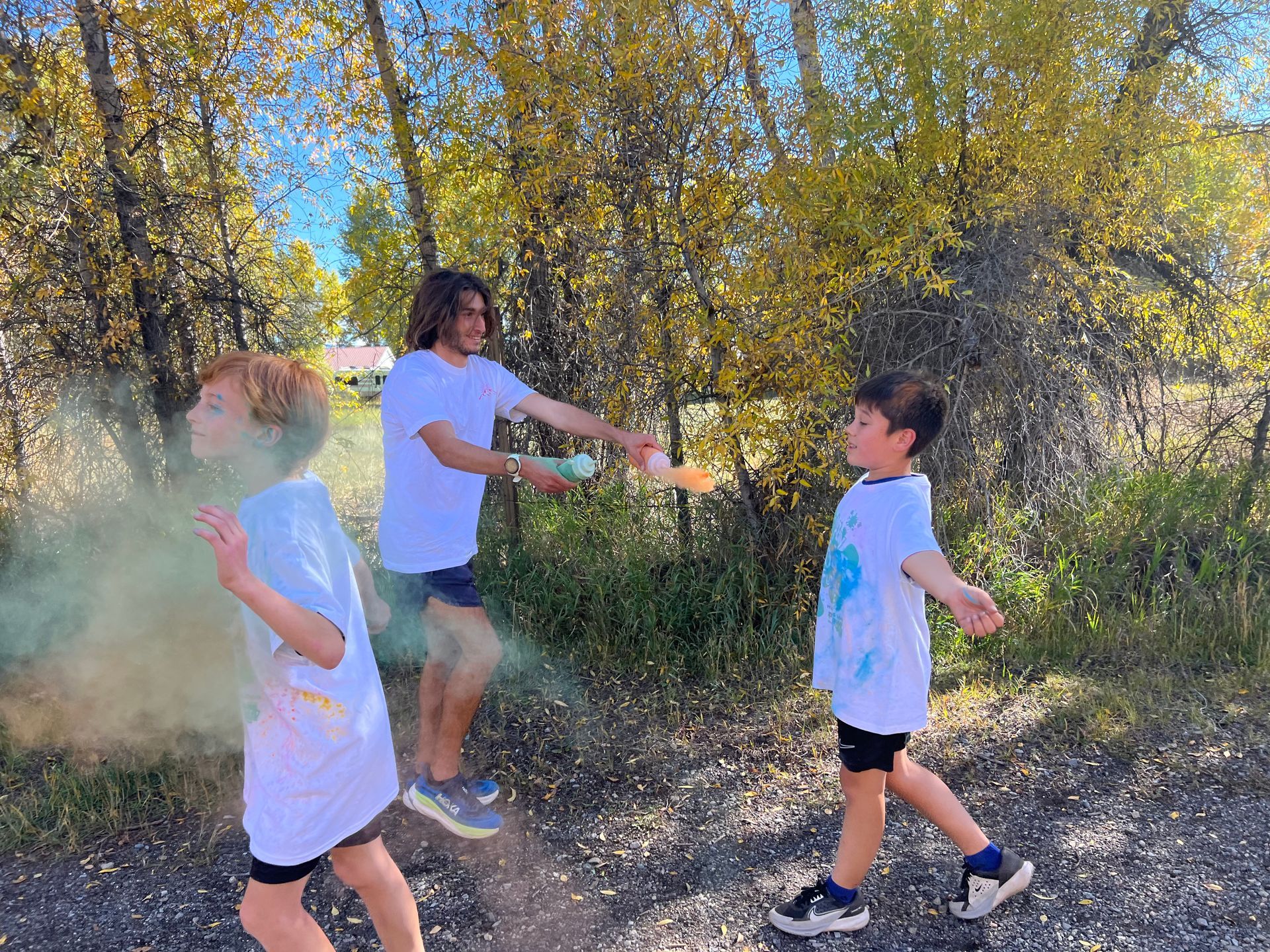 Three people throwing colored powder in a park; sunny day, trees in background, white t-shirts, green and yellow powder.