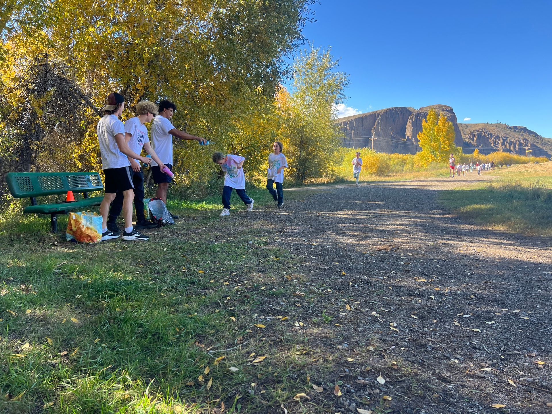 Group of people on a path near a bench, with trees and a mountain in the background. Sunny, outdoor setting.