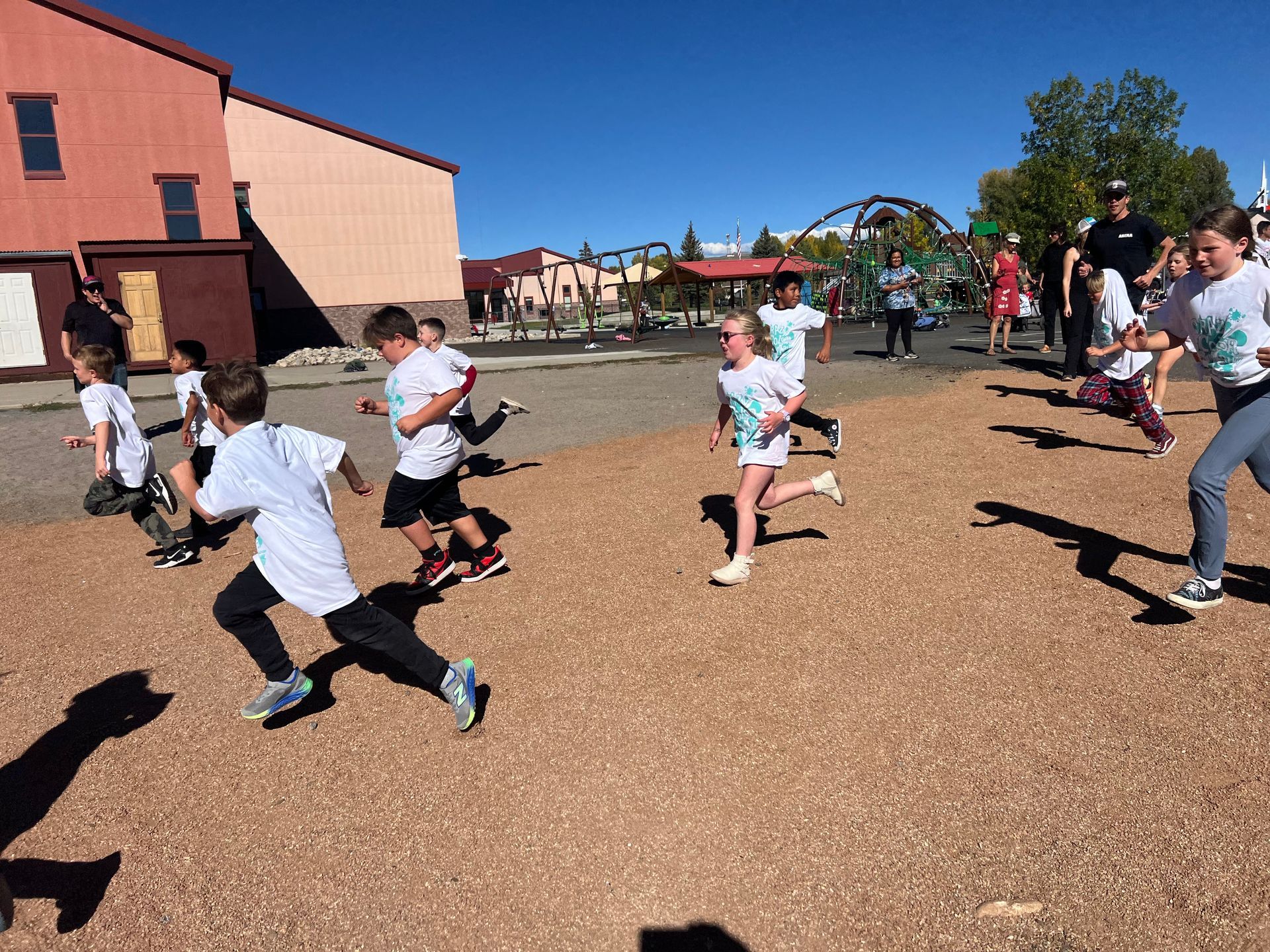 Children in white shirts running on a gravel surface near buildings and a roller coaster.