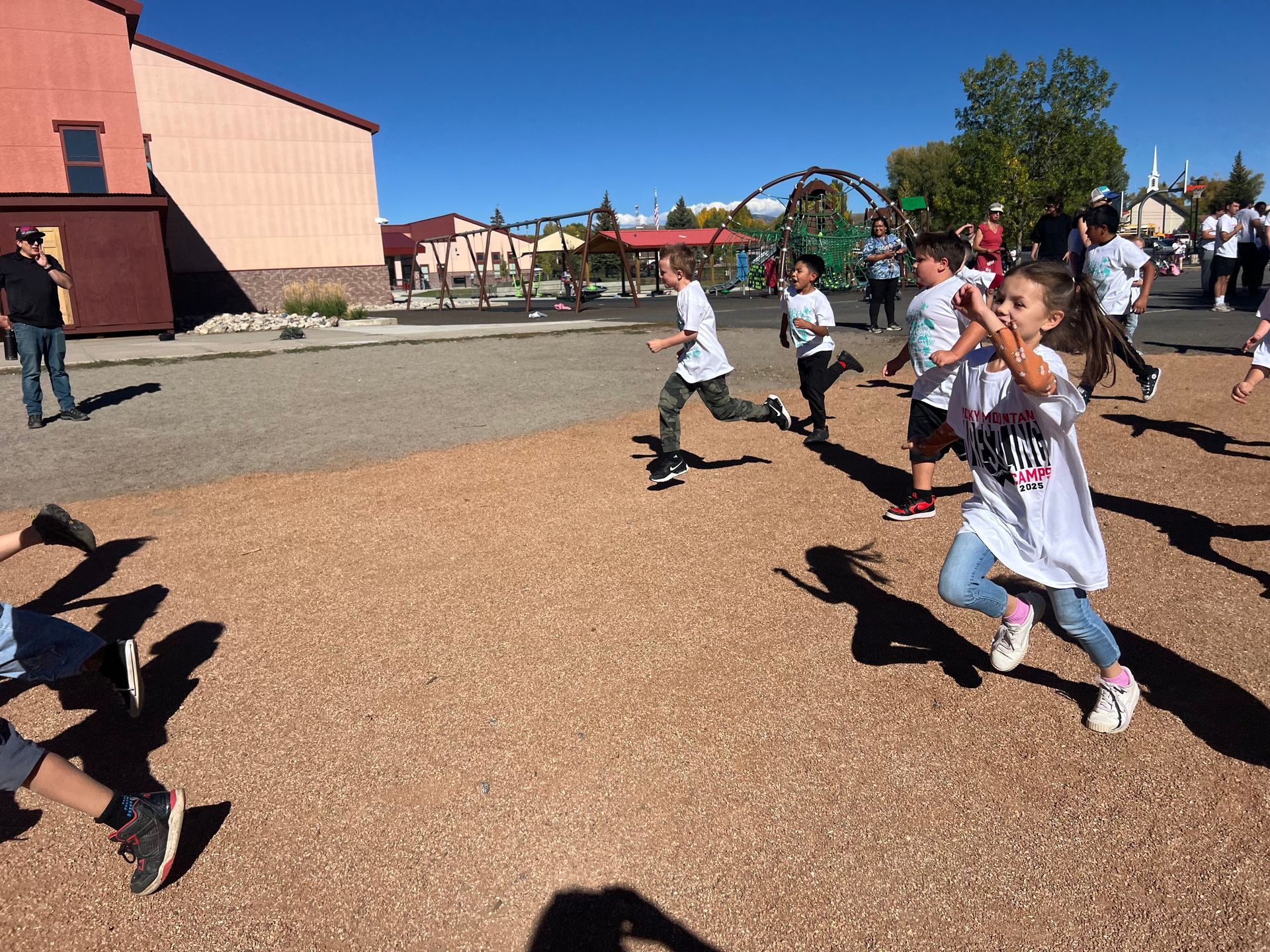Children running on a playground, wearing white shirts, sunny day. Brown ground, building in background.