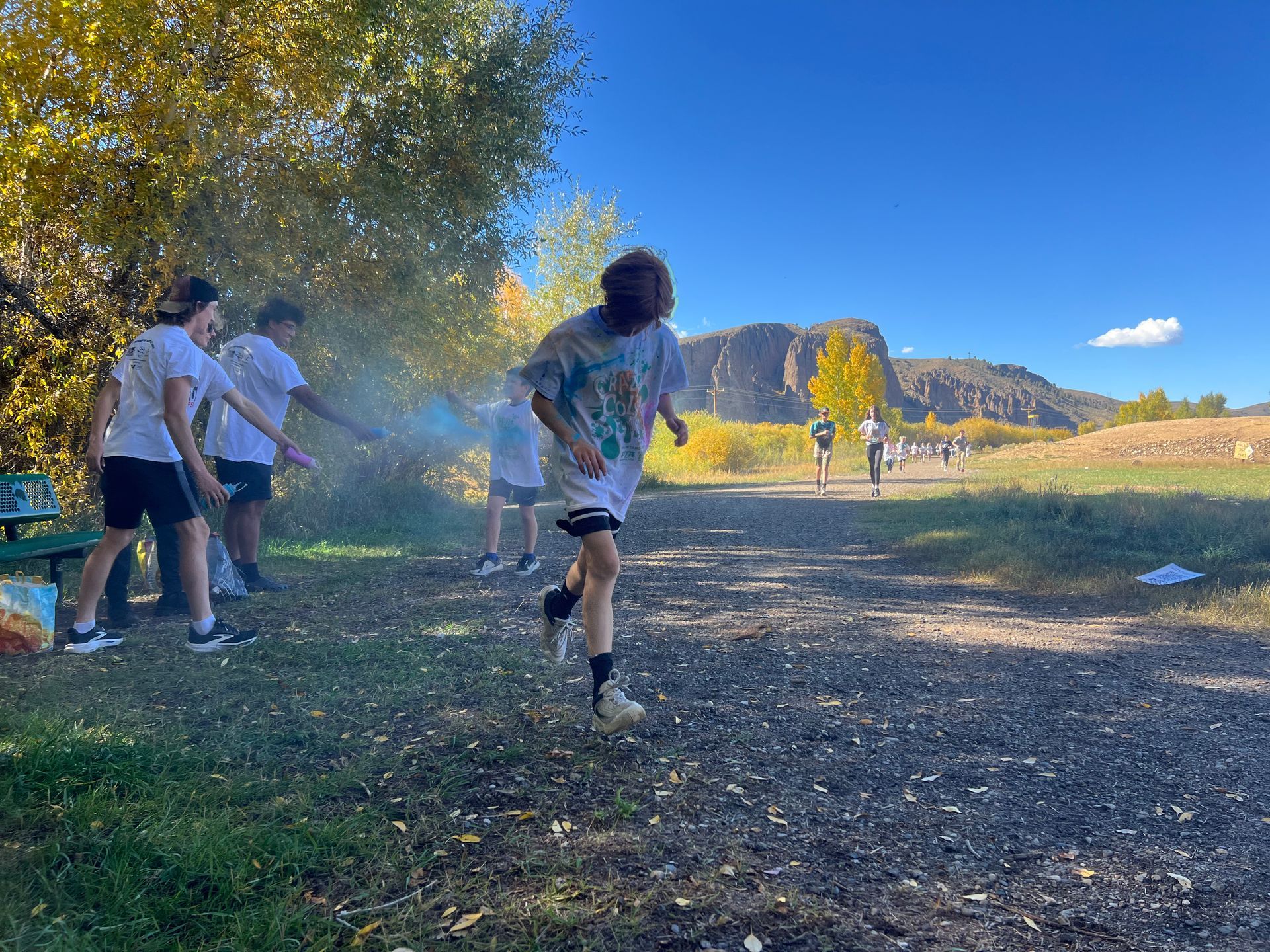 People covered in colored powder, running on a path outdoors with trees and a mountain in the background.