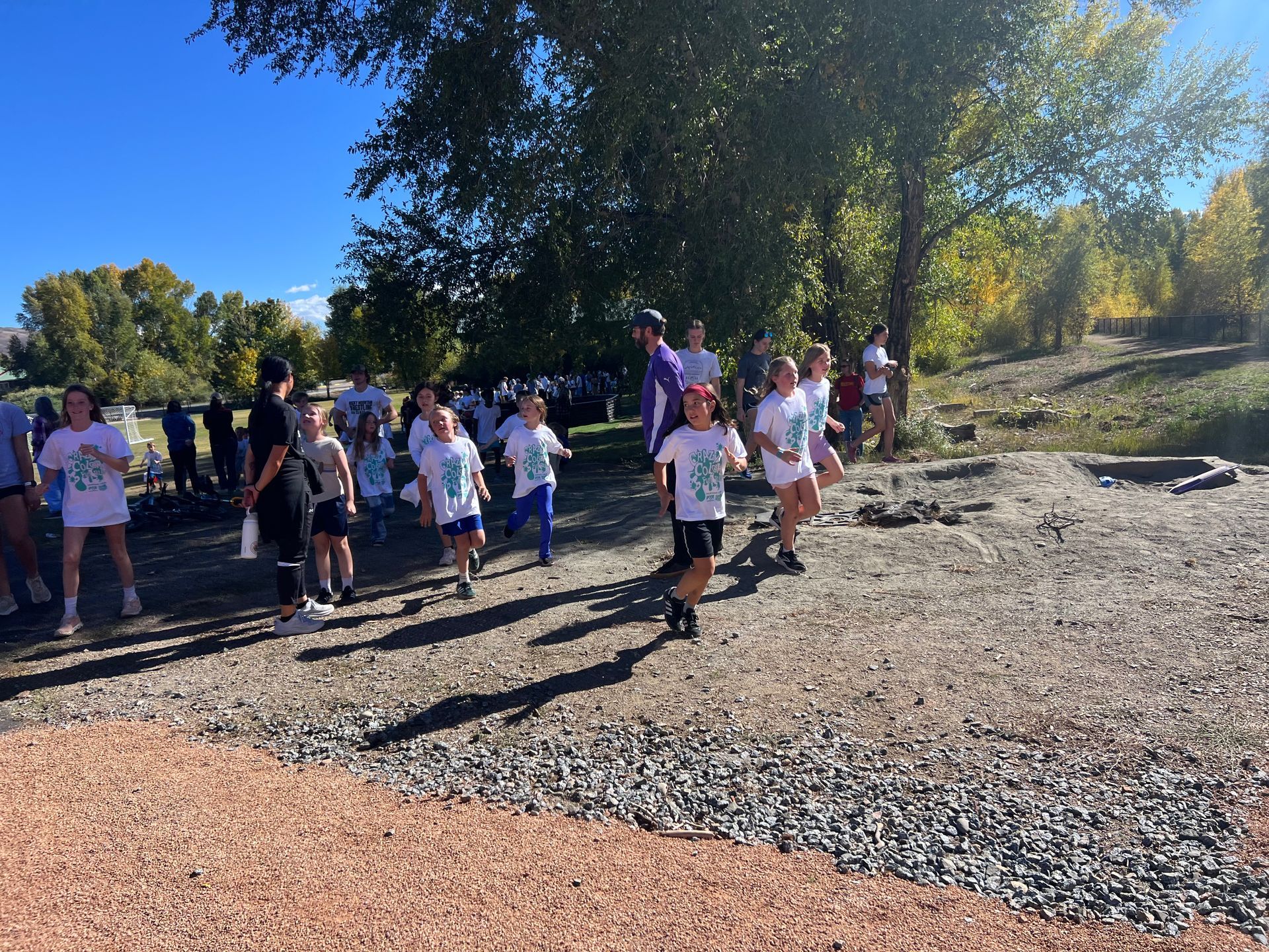 Runners in white shirts and shorts on a gravel path, trees in the background, sunny day.