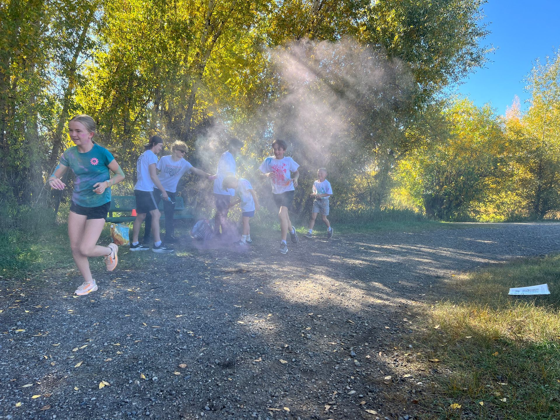 Runners passing through a water spray on a gravel path near trees on a sunny day.