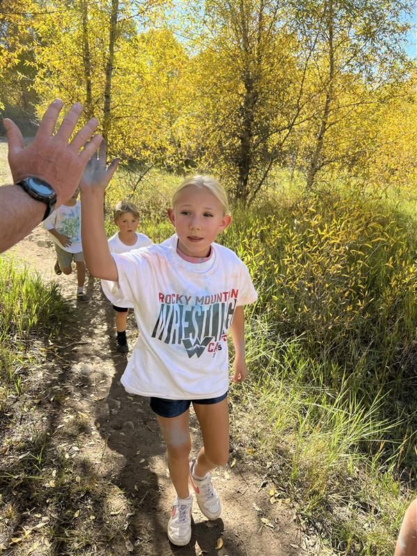 Girl giving a high-five, muddy, wearing a white shirt and shorts, running outdoors with autumn foliage.