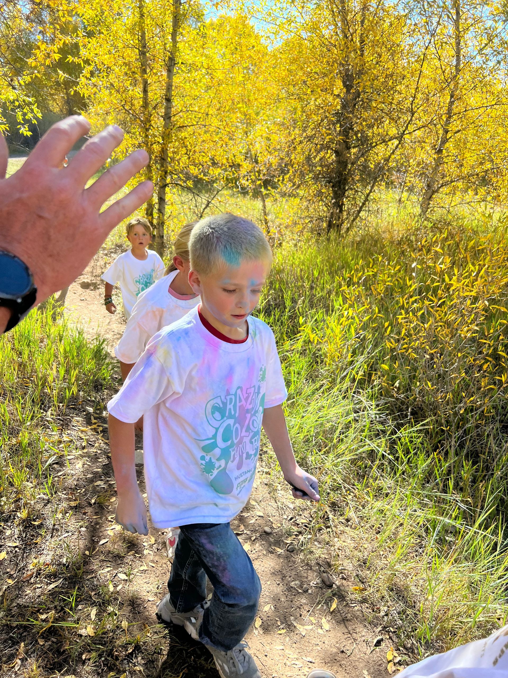 Boy with colorful powder on white shirt walks on a trail, with trees in the background.