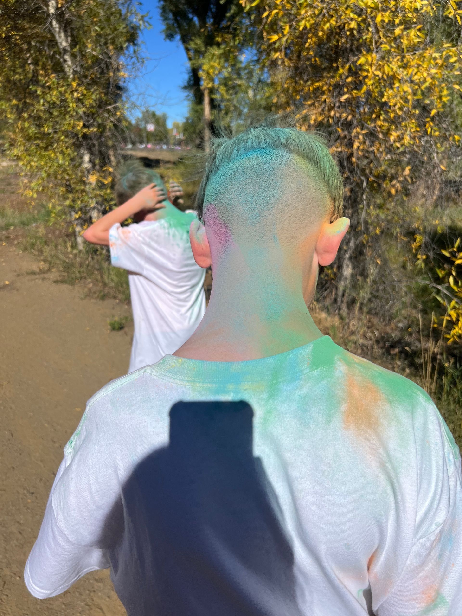 Person with colorful powder in hair and on shirt, outdoors with another person.