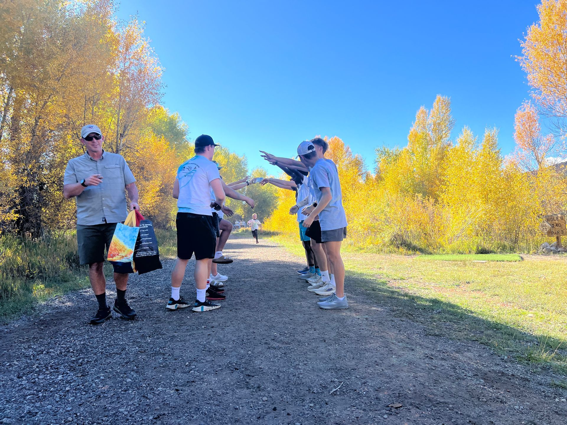 People high-fiving a runner on a gravel path lined with yellow fall trees under a blue sky.