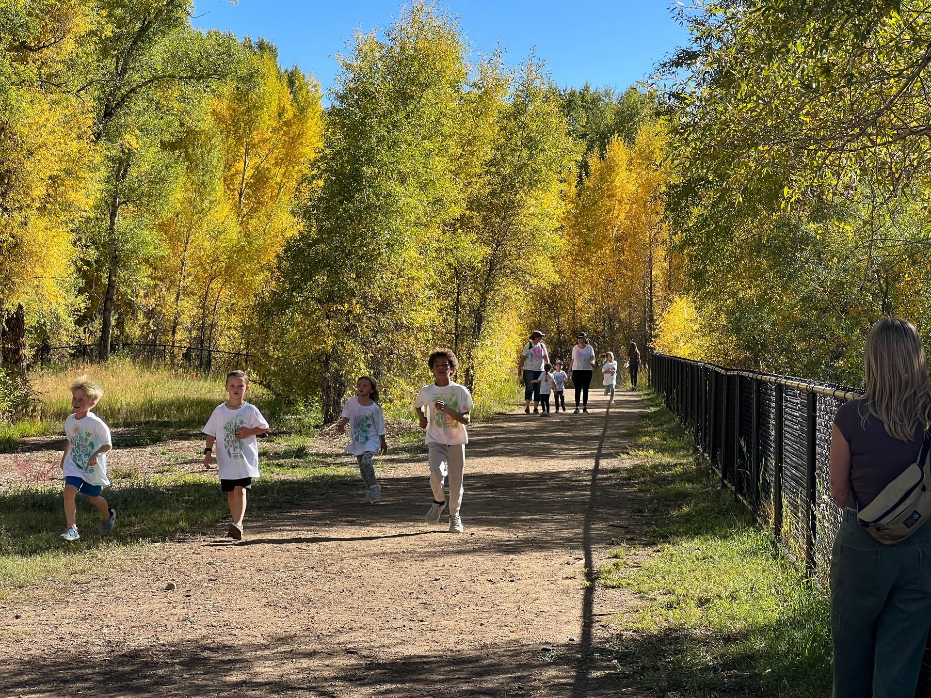 Children running on a dirt path alongside a black fence, surrounded by fall foliage. Sunny day.