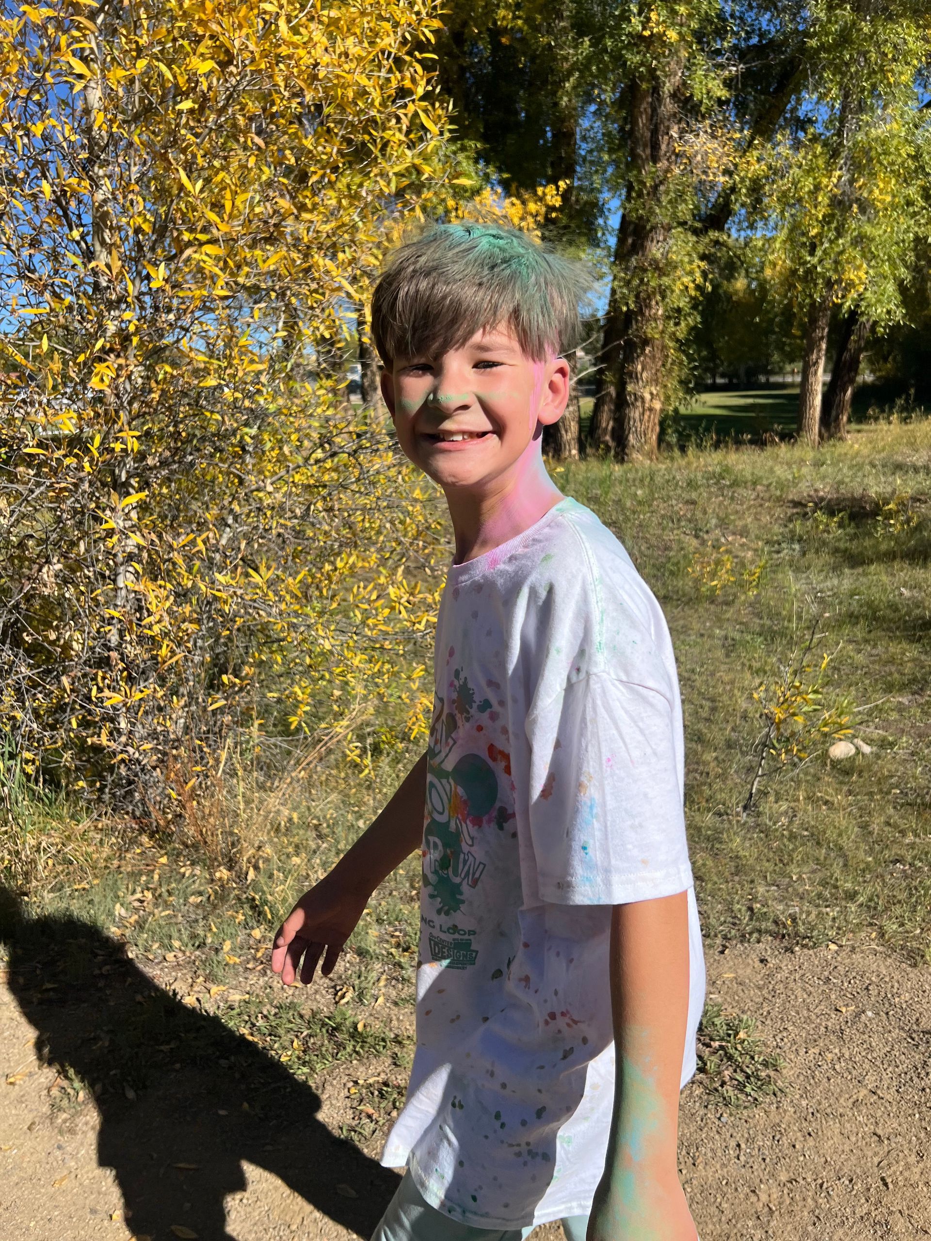 Boy smiling, covered in colorful powder. Outdoors with autumn foliage.