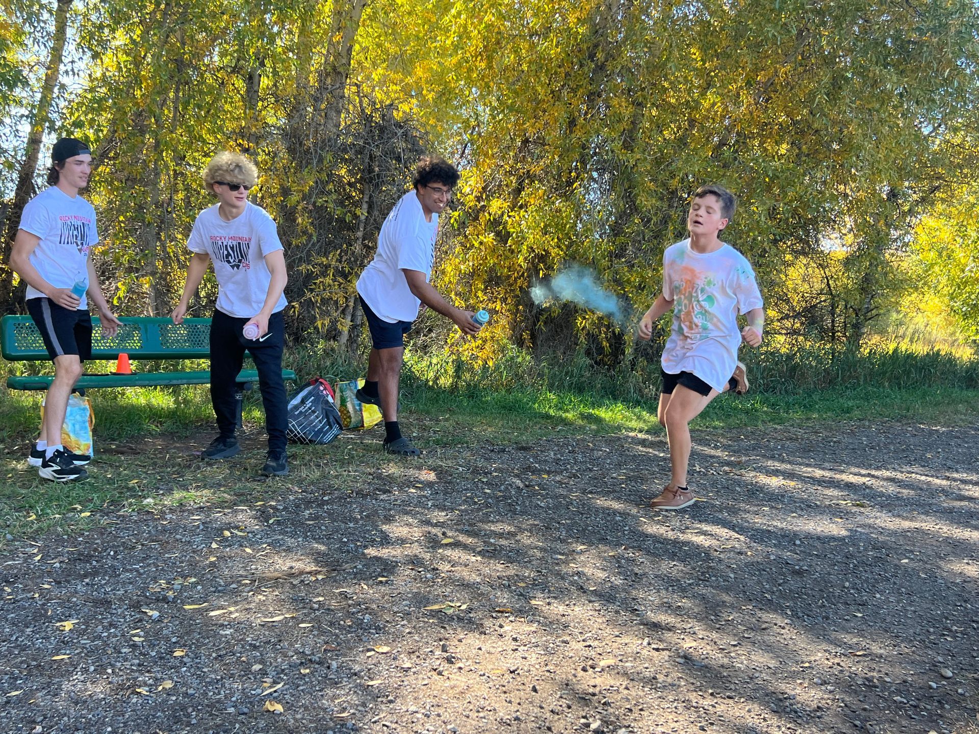 Four people in white shirts and shorts are throwing colored powder. They are outside on a fall day.