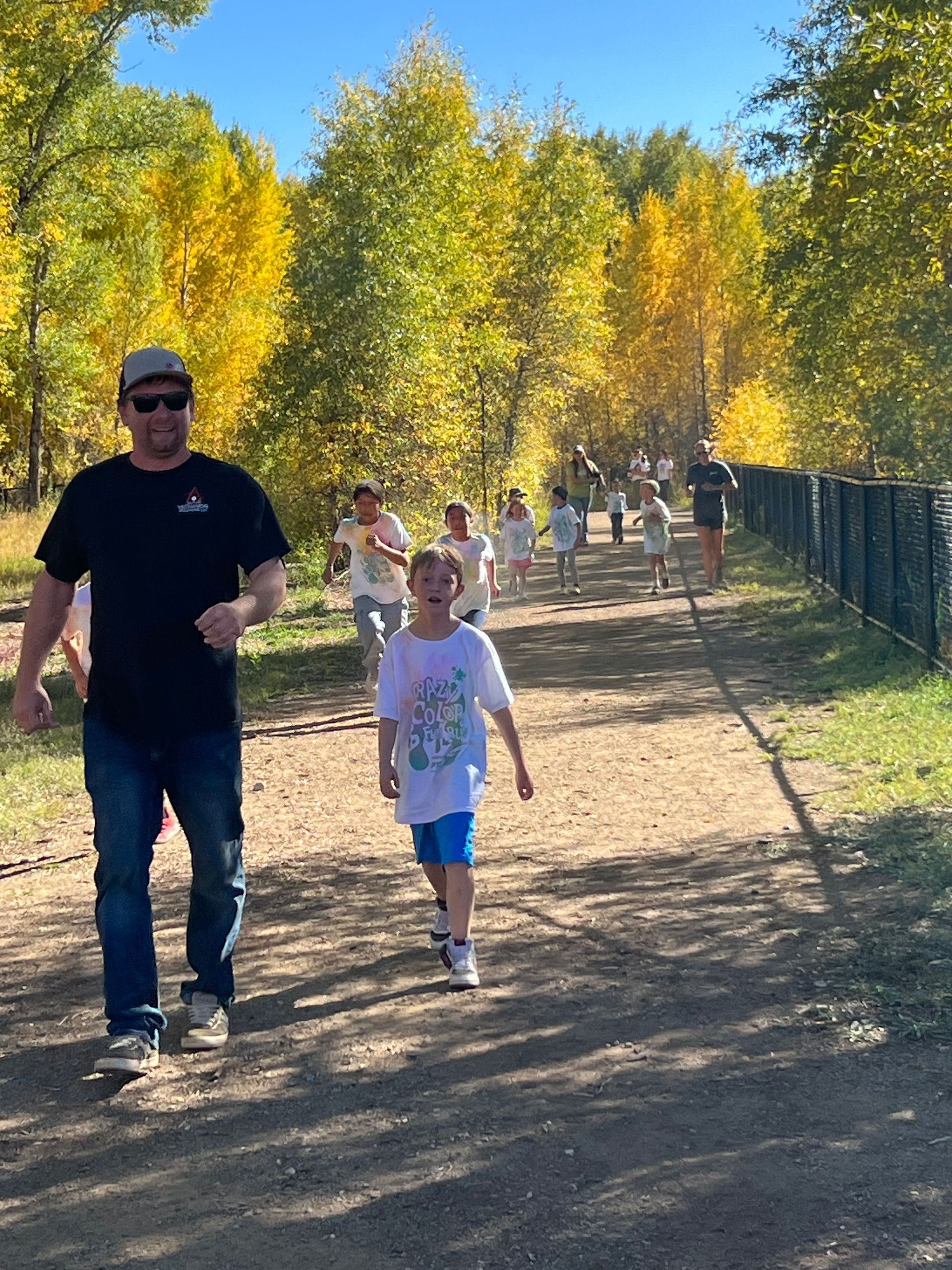 A group of people running on a dirt path next to a fence, trees with yellow leaves in background.
