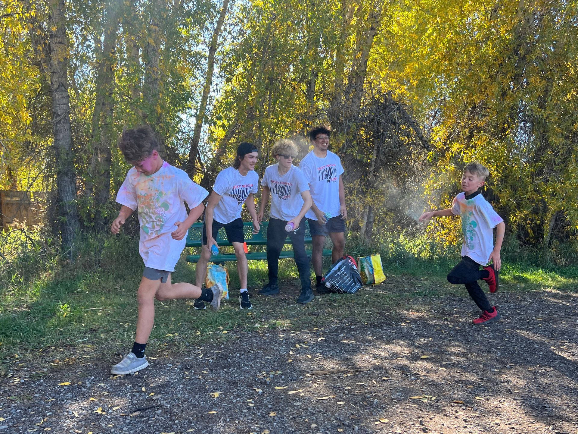 Kids covered in colorful powder running near a park bench, trees with autumn leaves in the background.