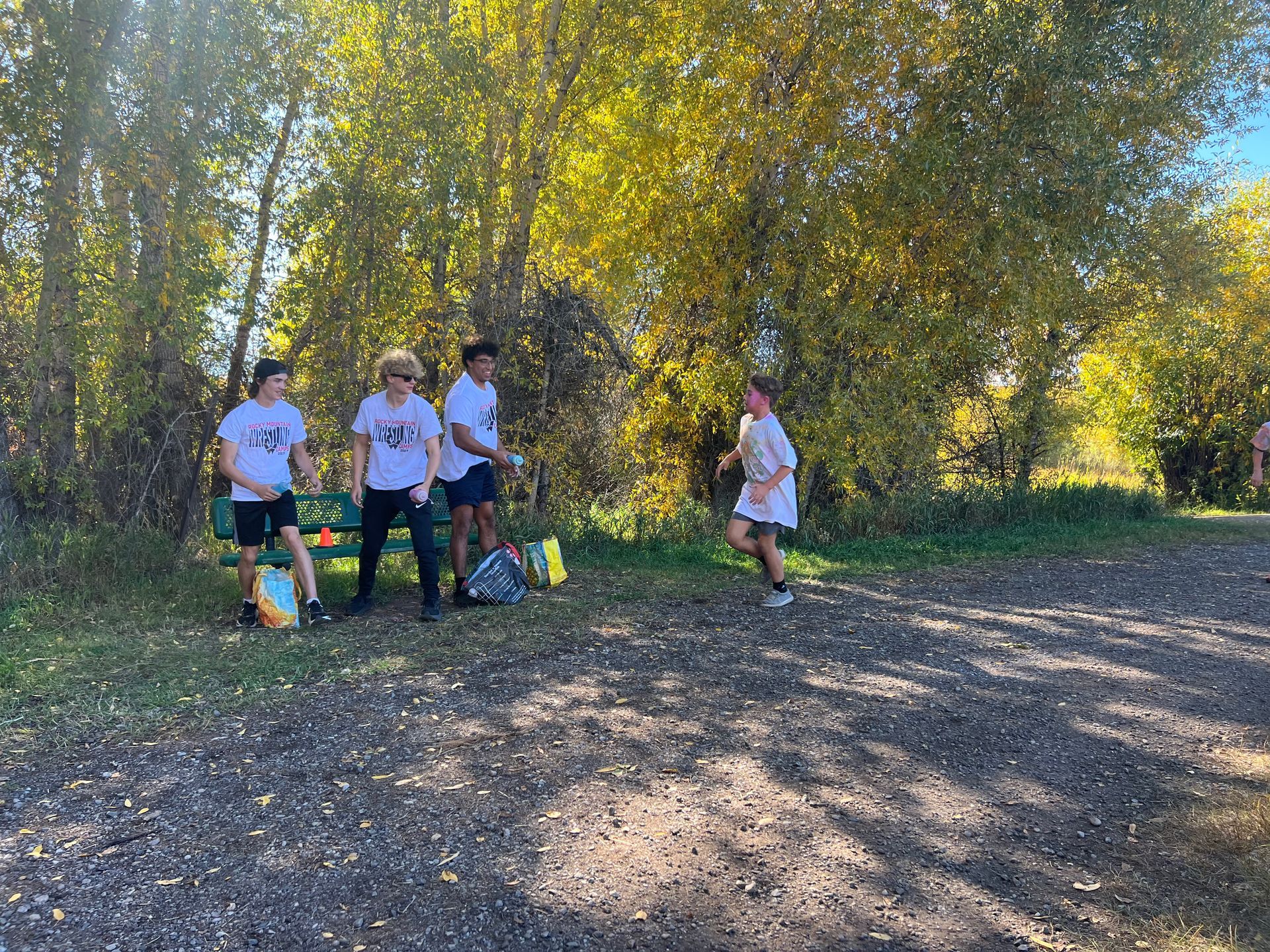 Group of people near a bench and trees, some wearing white shirts and shorts. One person is running. Autumn setting.