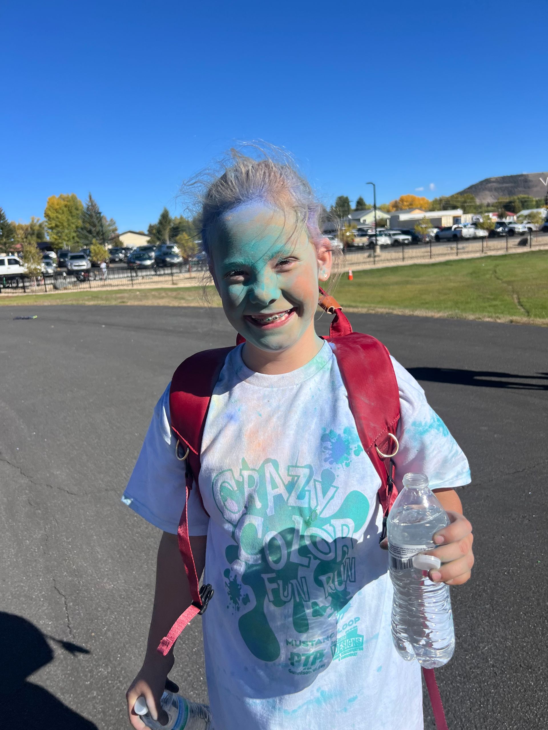Girl covered in colorful powder smiles, holding water bottle, outdoors.