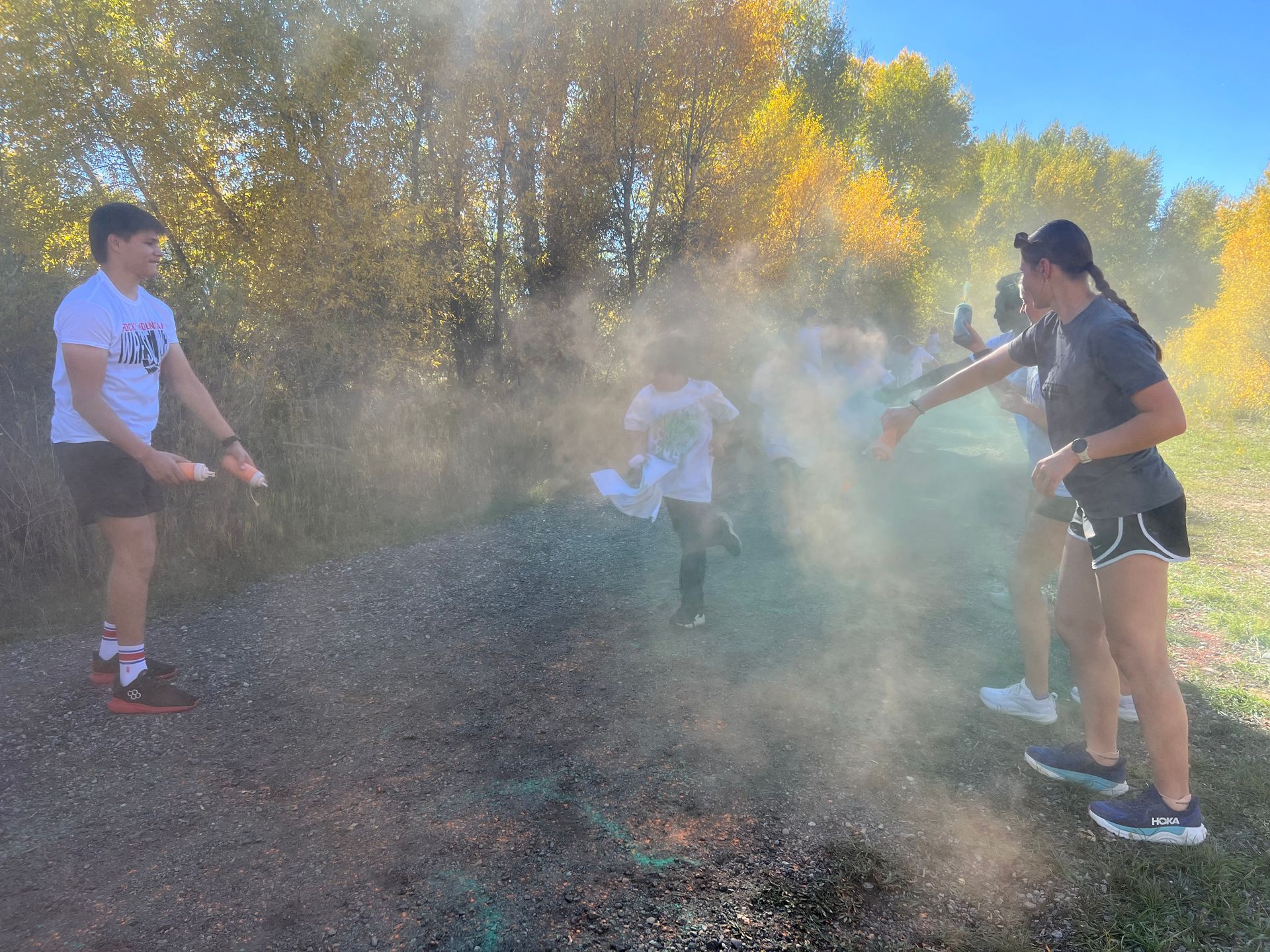 People throwing colored powder as runners pass on a trail. Sunny day with fall foliage.