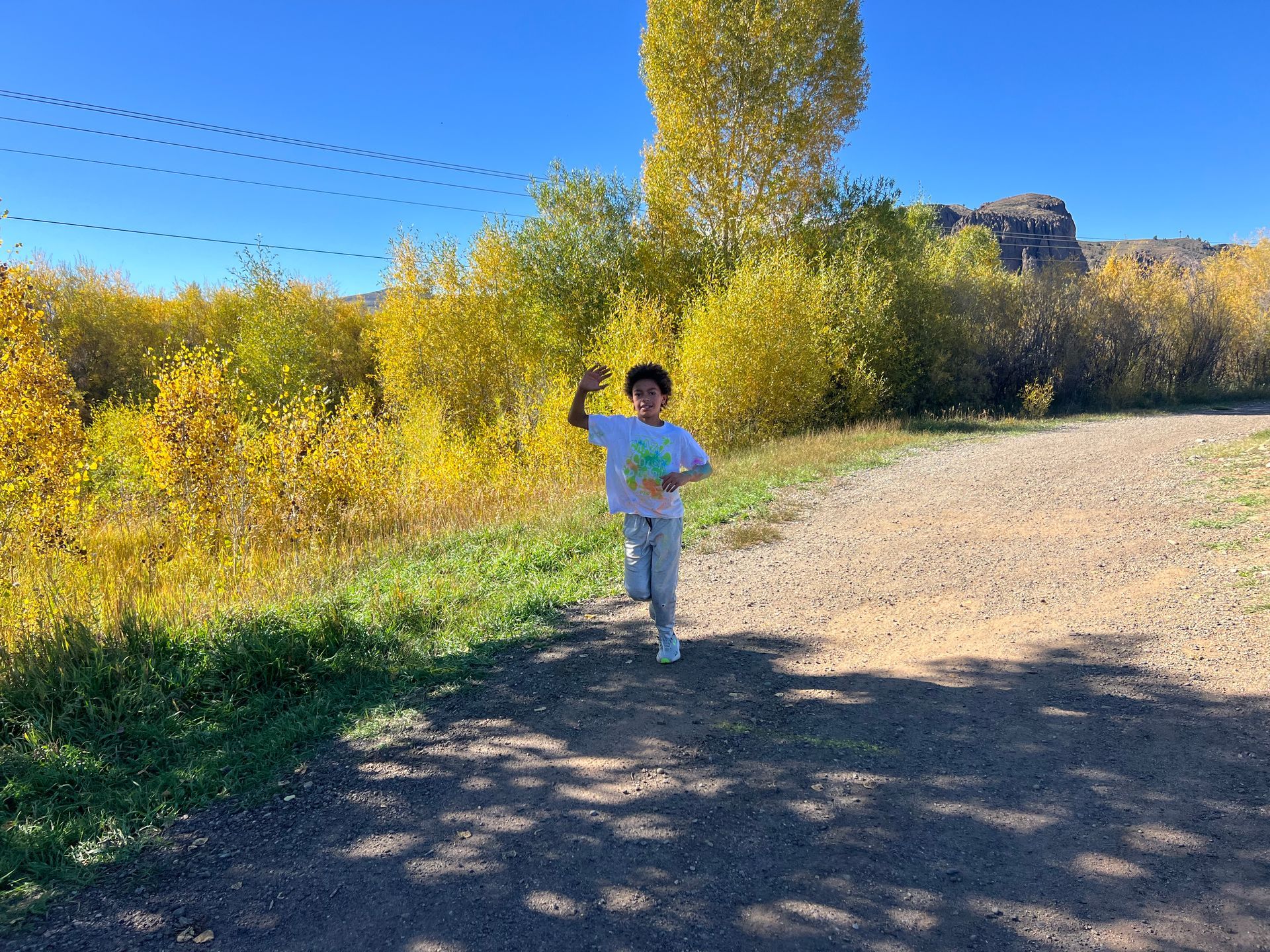 Young person running on a dirt path, raising a hand. Surrounded by yellow trees on a sunny day.