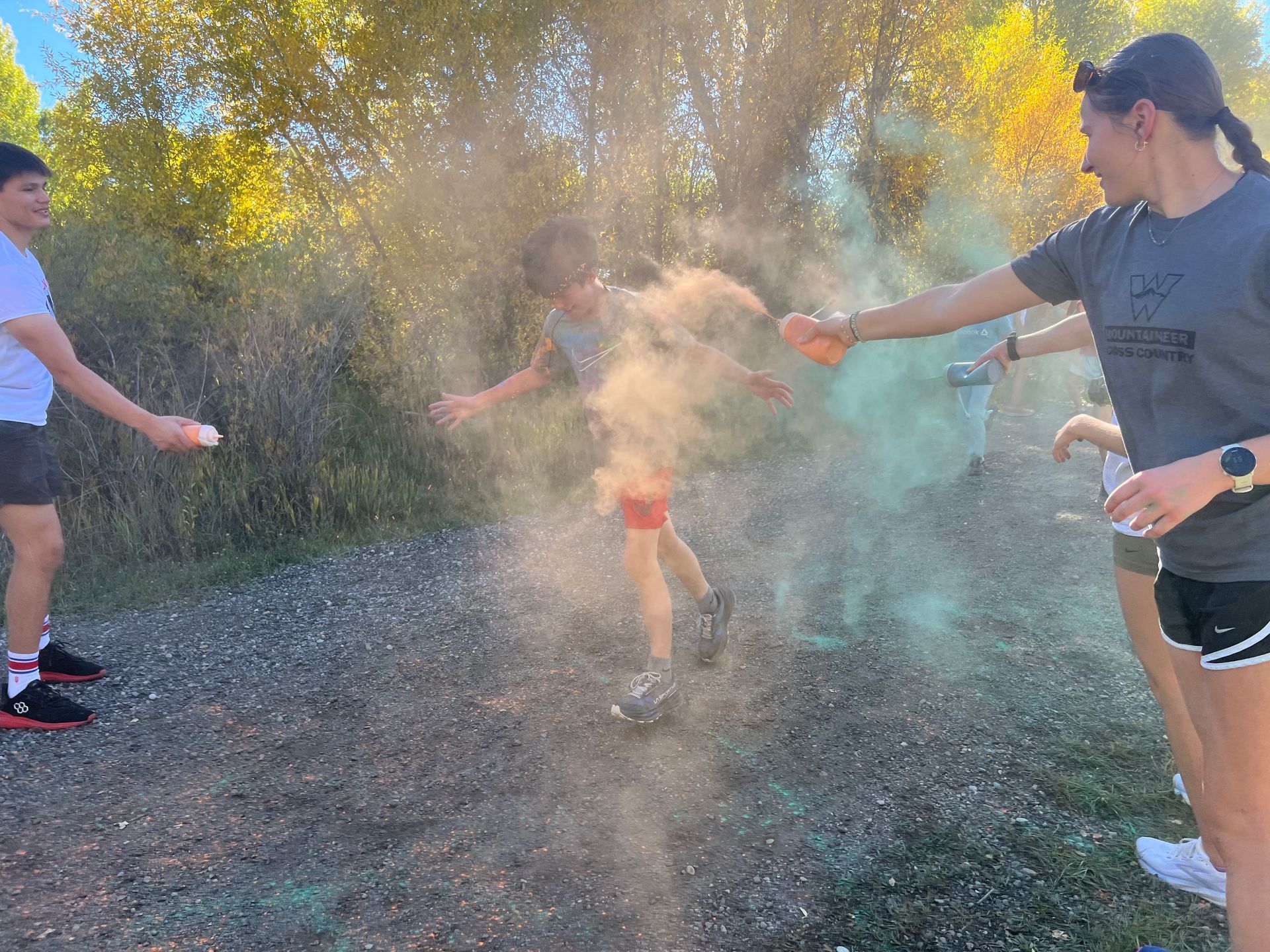 People spraying colored powder on a person running. Outdoors with trees and gravel path.