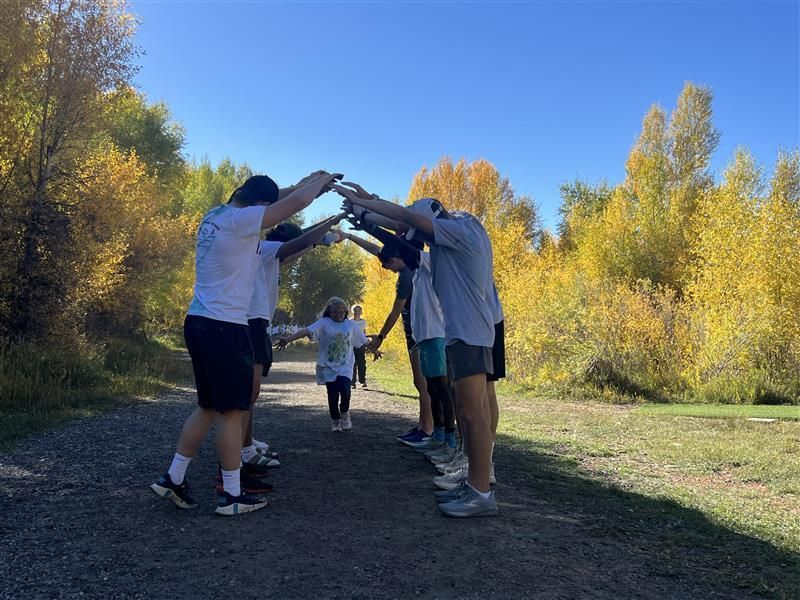People making an arch with their arms for a child running on a path, surrounded by yellow trees under a blue sky.