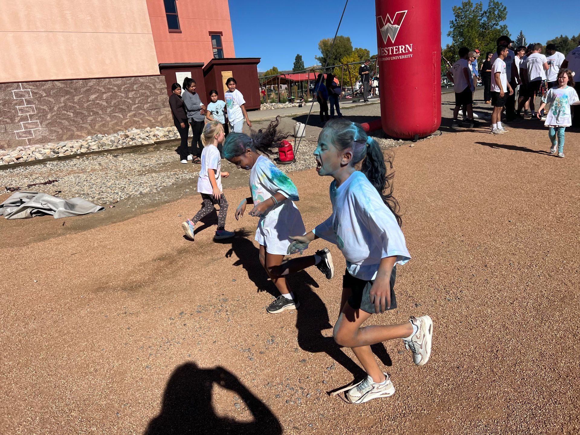 Children running toward a red inflatable arch, covered in colorful powder, at an outdoor event.
