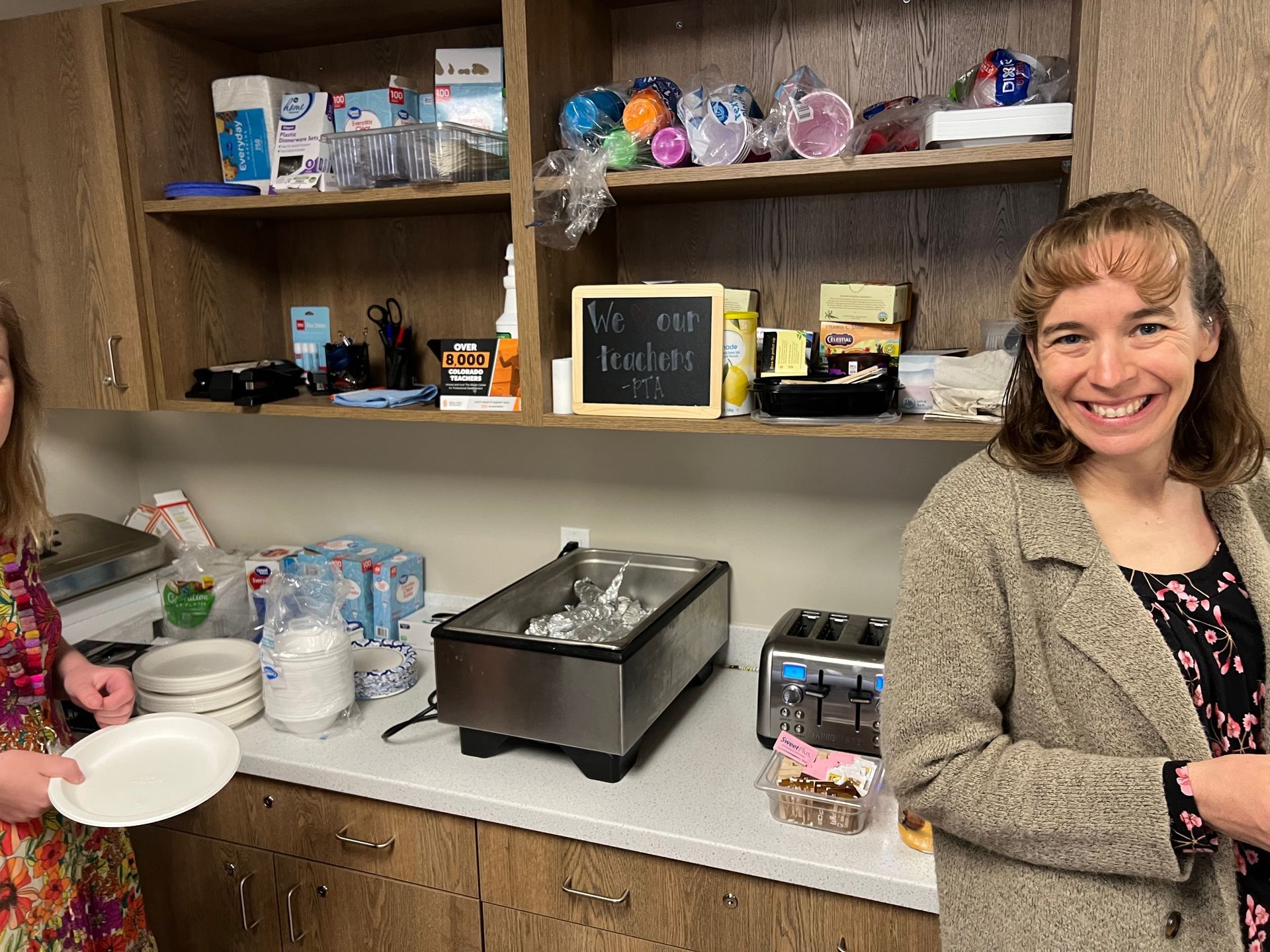 Two women in a kitchen. One holds a plate; the other smiles. Countertop with toaster, food warmer, and supplies. Shelves above.