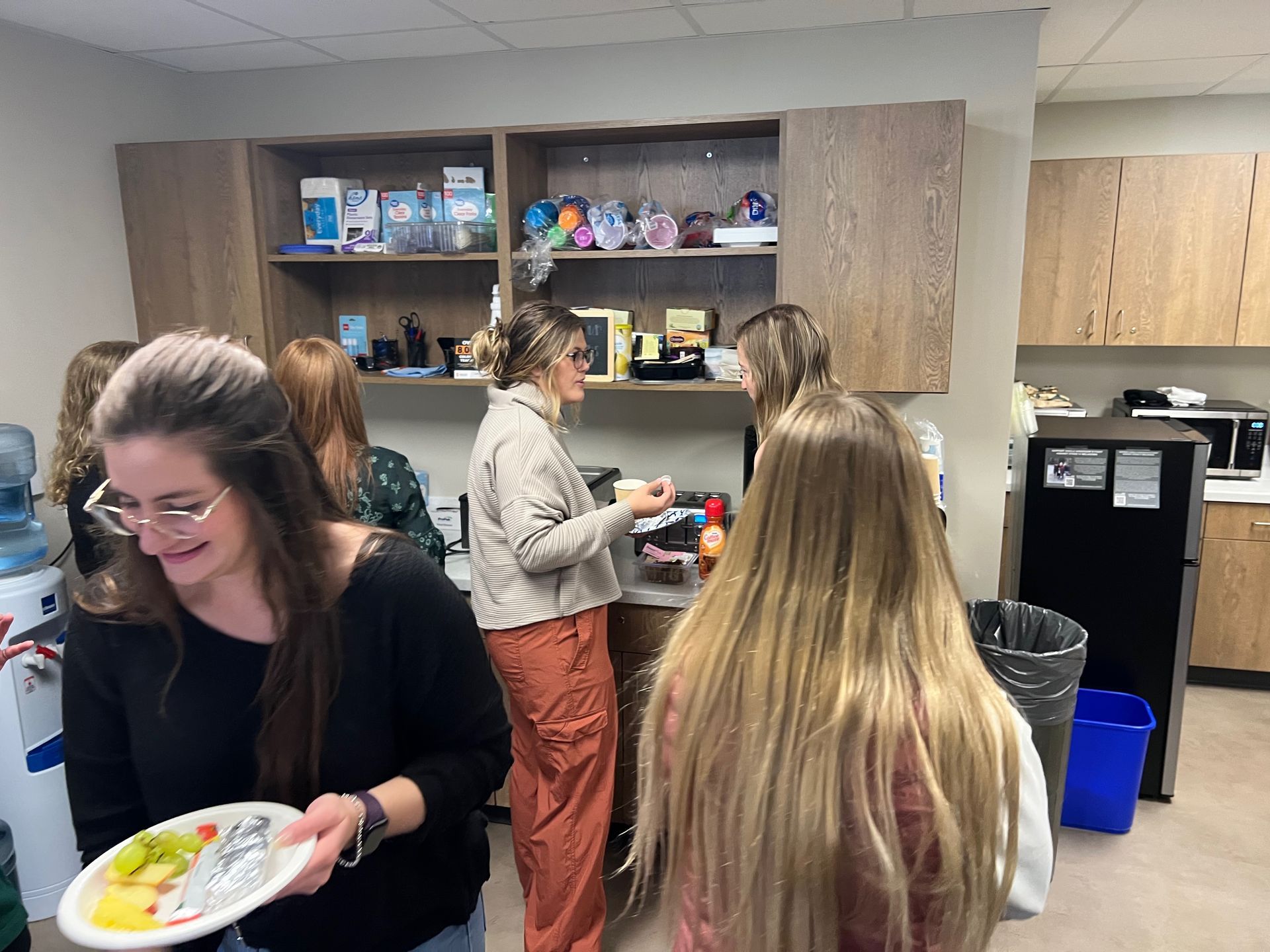 People in an office kitchen, some holding food and drinks, near coffee station and cabinets.