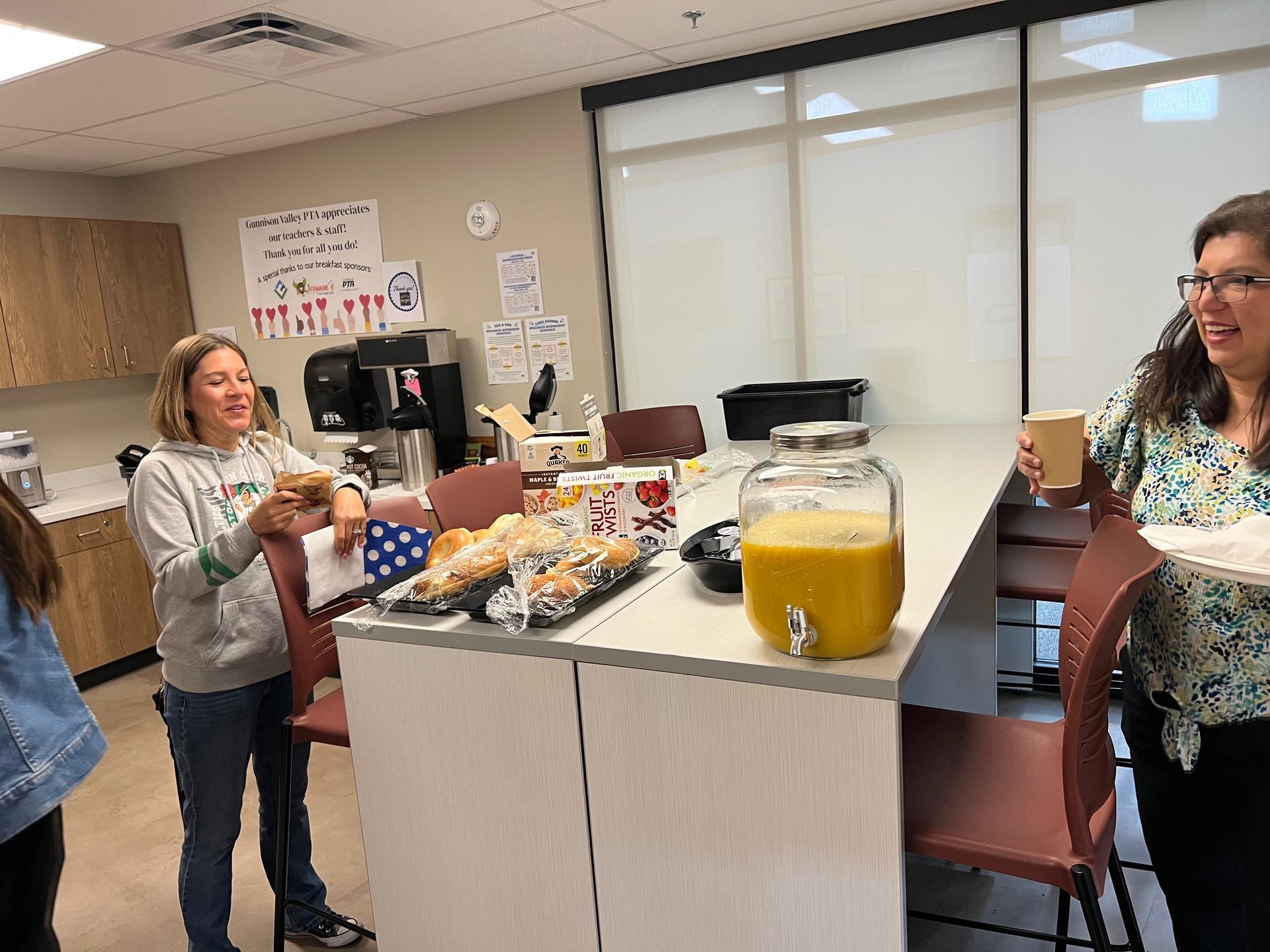 People at a break area with food and drink. One woman holding a drink, another standing near the counter.