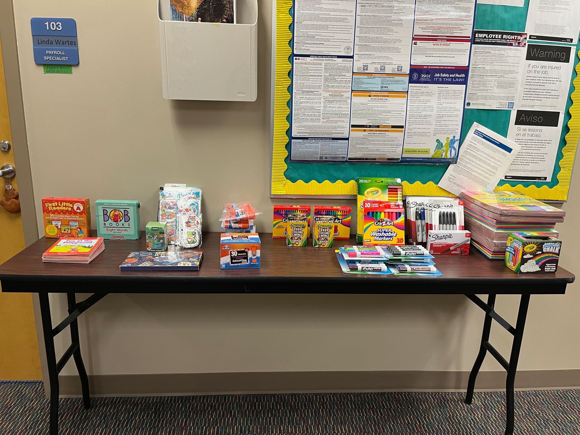 A table displaying various boxed snacks and school supplies in front of a bulletin board.