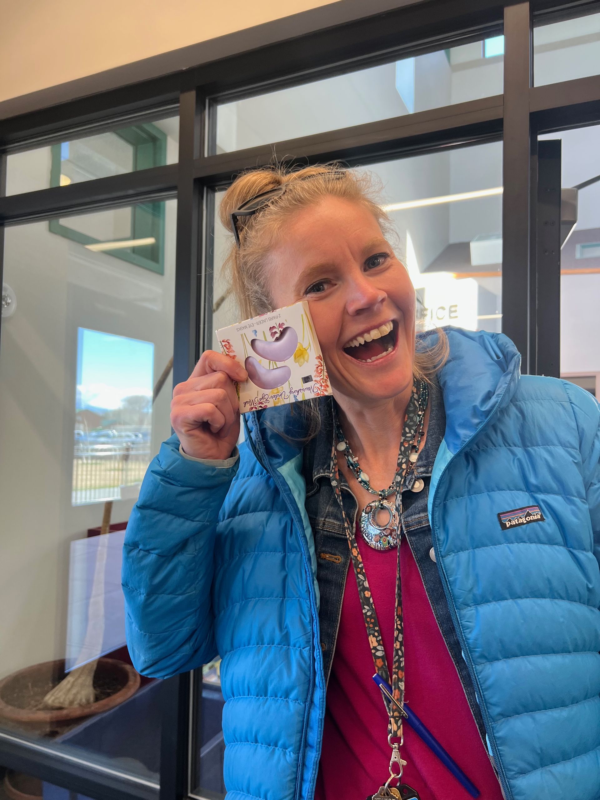 Woman with blonde hair smiling, holding a card with a cartoon character; wearing blue jacket and necklace.