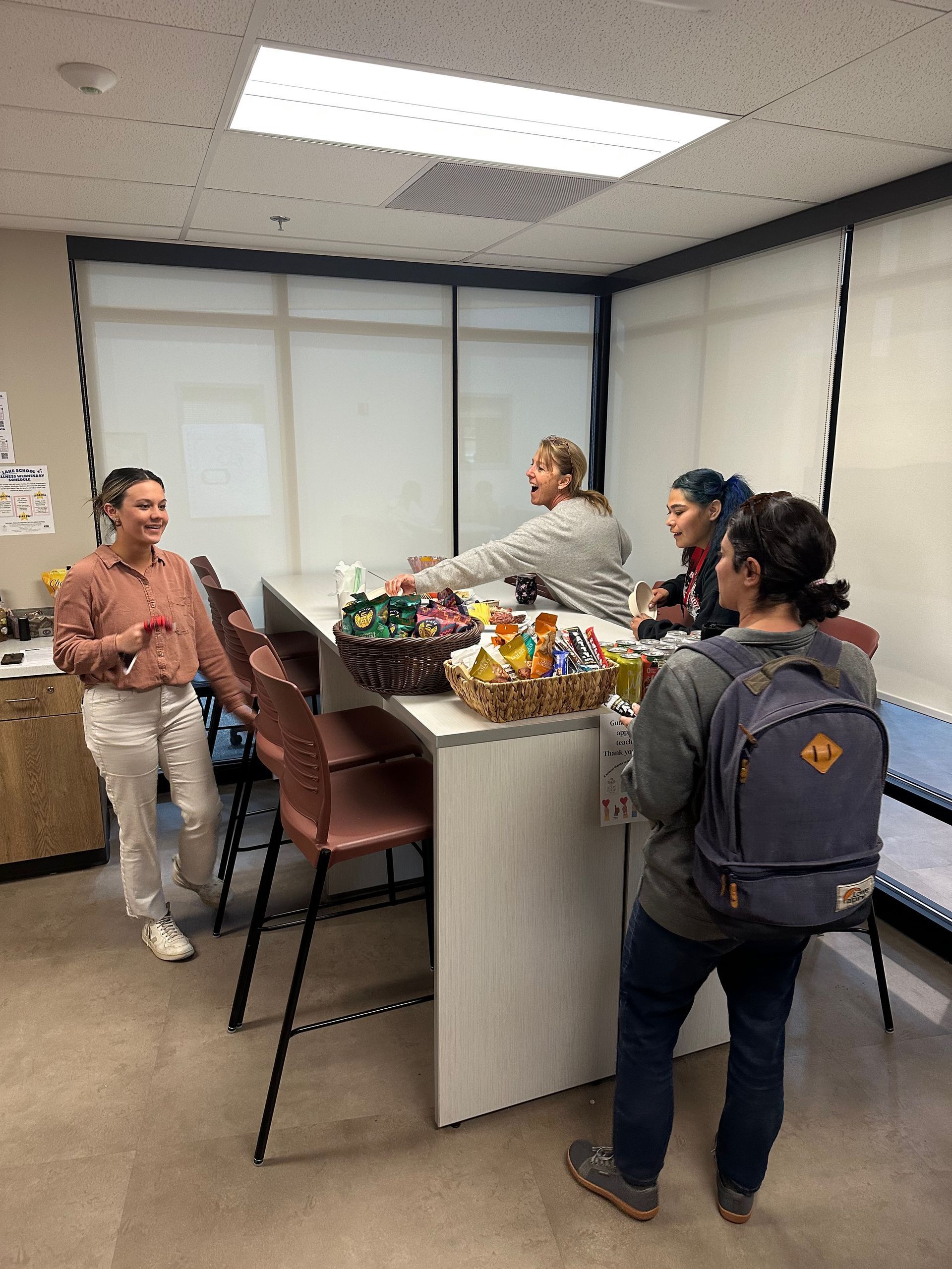 Four people at a table with snacks, in a brightly lit office setting. One person speaks, others look on.