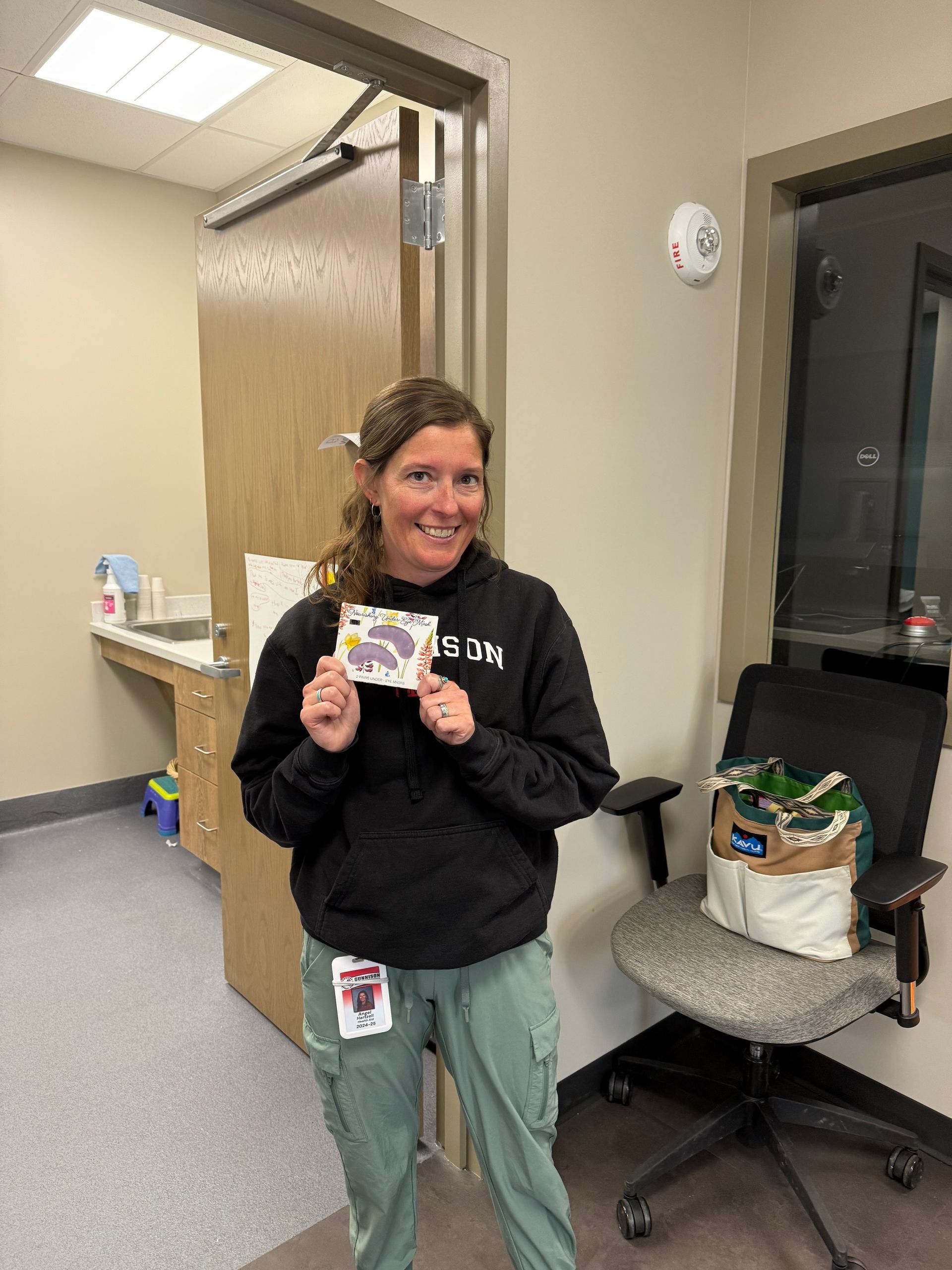 Woman in green pants and black hoodie holds a card, smiles, stands in a clinic room near a chair and groceries.
