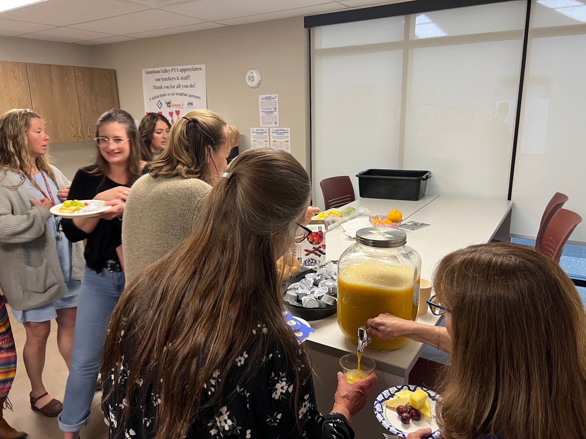 People at a gathering, serving juice and fruit near a table. White walls, windows, and casual attire.