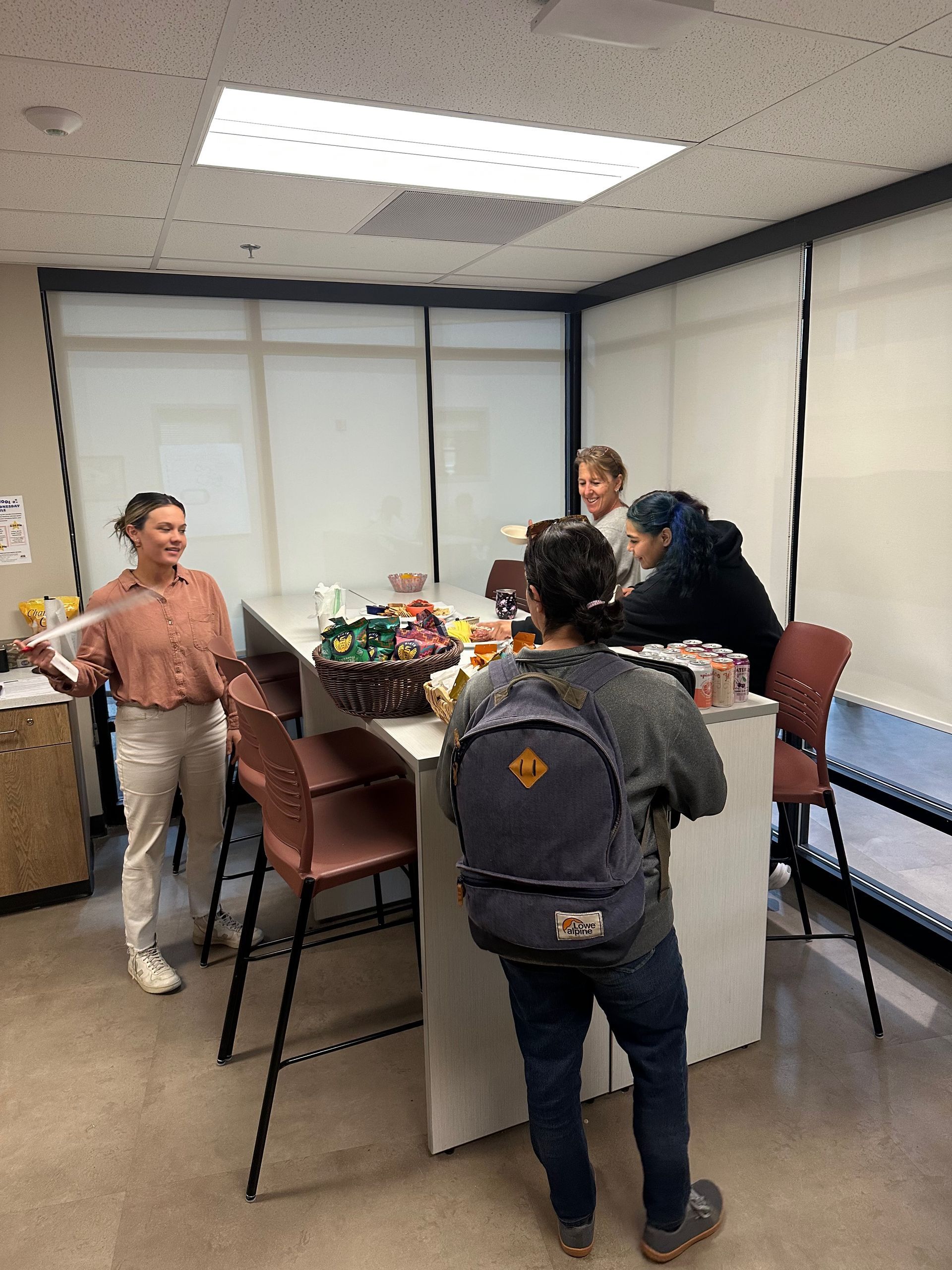 Group of people gathered around a counter with snacks in an office. One person is holding papers, and another has a backpack.