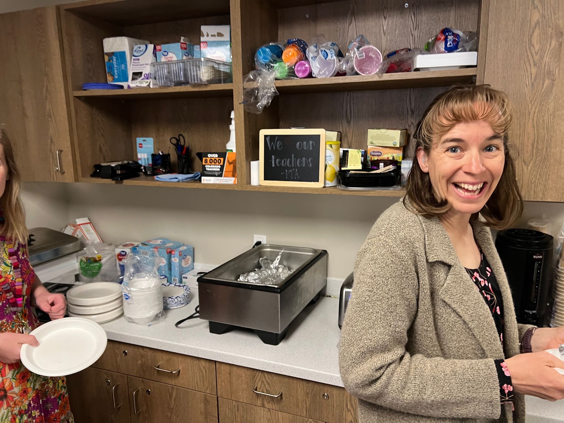 Two women in a breakroom. One smiles near counter with plates and food warmer. Shelves hold supplies.